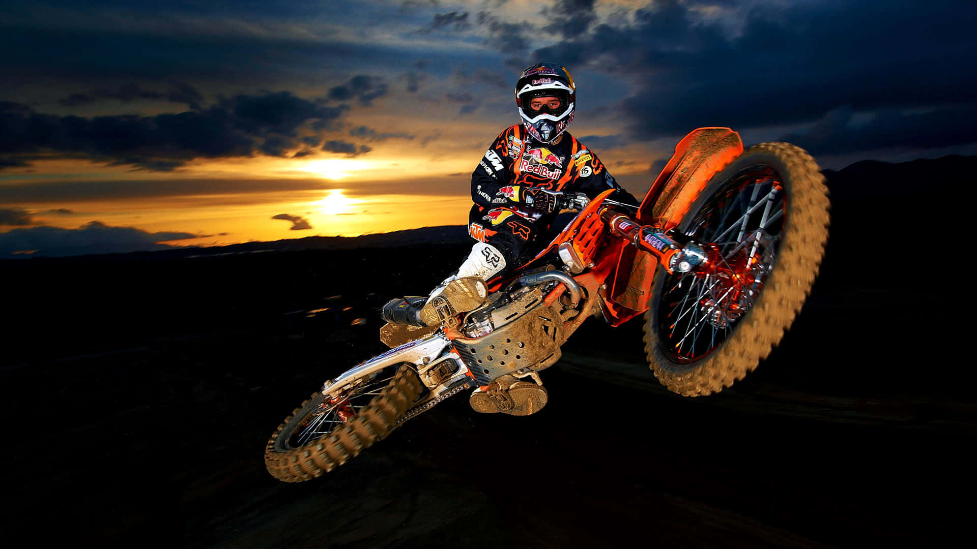 Motocross 桌布 壁紙 — 一名全副裝備的越野機車手在金色雲彩的戲劇性日落天空下表演大膽的後輪平衡特技。