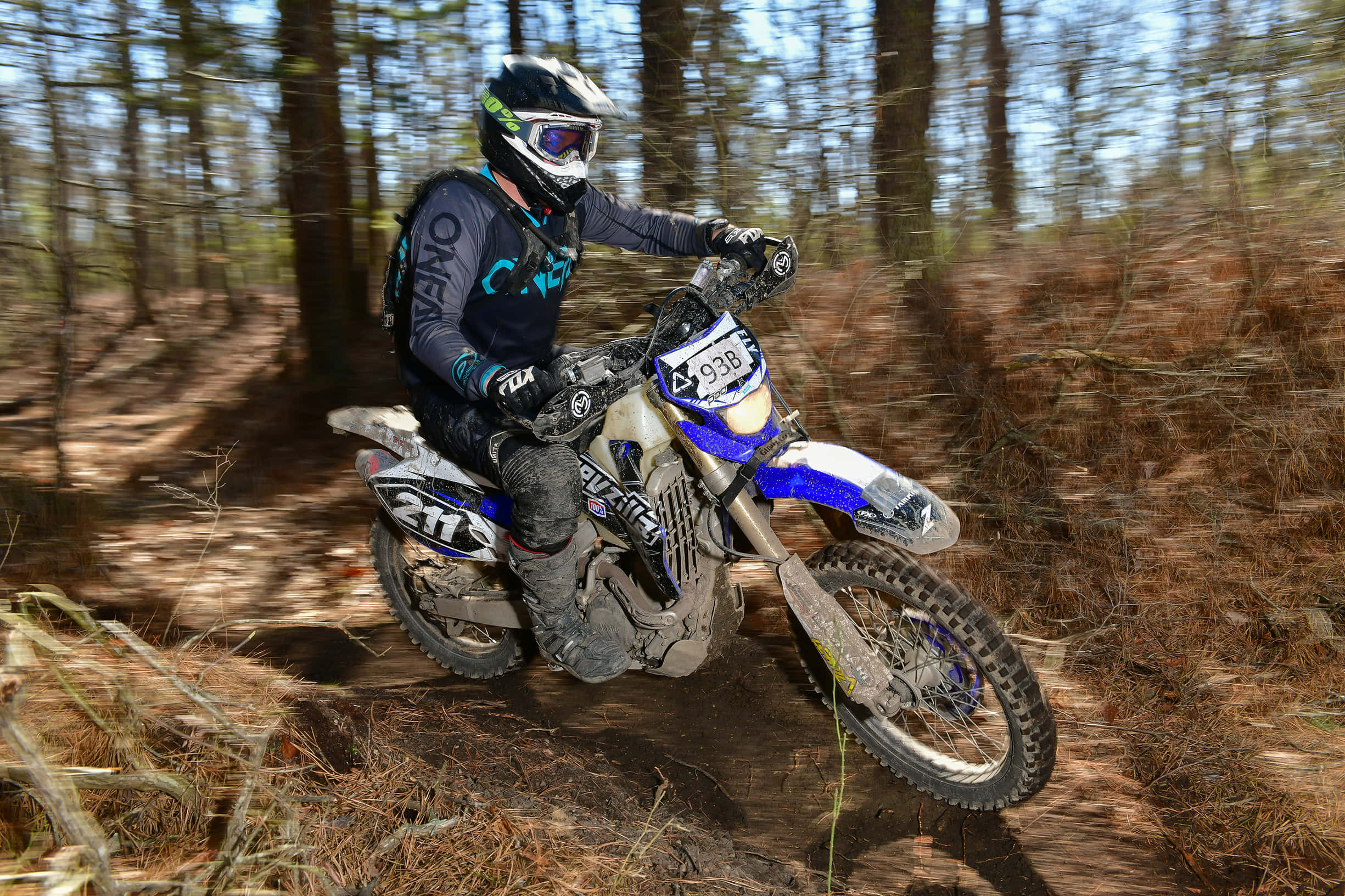 Dirtbike 桌布 壁紙 — 一名全副裝備的越野機車騎士，在陽光明媚的森林小徑上，騎著藍白相間的泥地機車奔馳，揚起塵土。