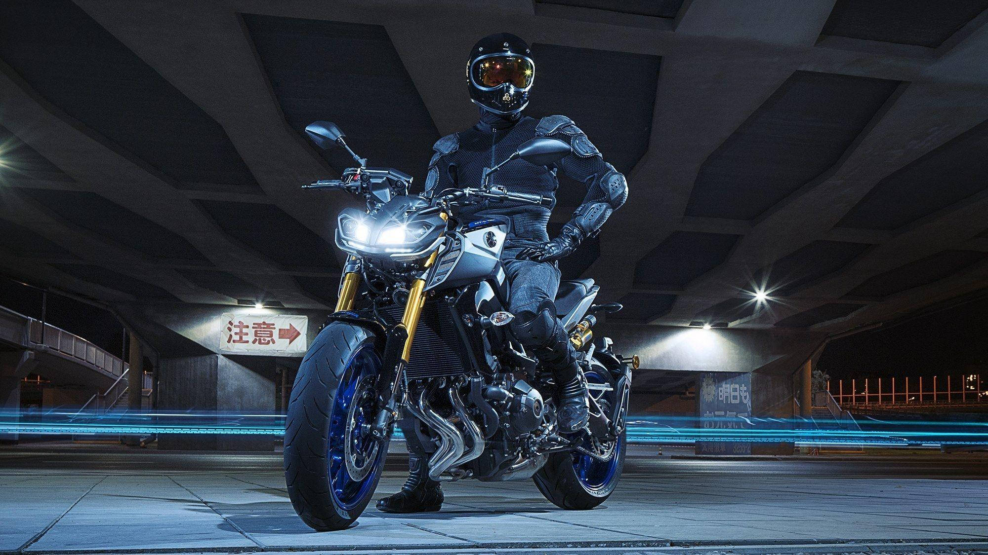 yamaha mt 15 桌布 壁紙 — 一名全副裝備的騎士在夜間站在幾何混凝土天橋下的時尚機車上，伴隨著藍色光條。