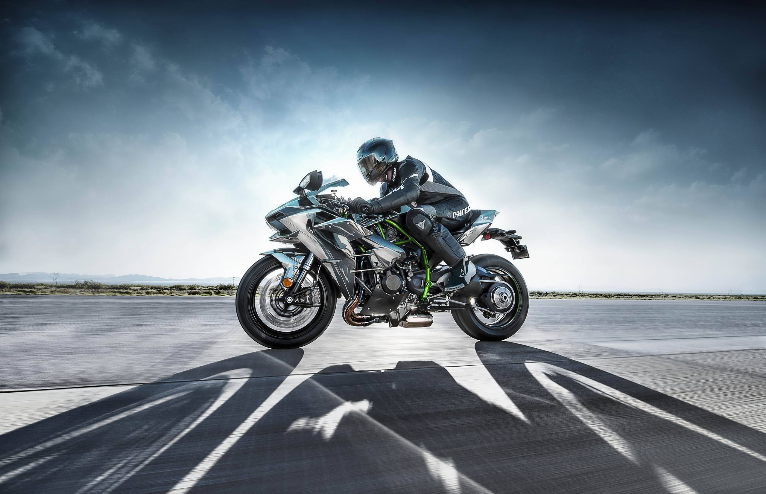 Kawasaki H2r 桌布 壁紙 — 一名全副裝備的騎士在戲劇性的多雲天空下，騎著一輛時尚的銀綠色運動摩托車傾斜入彎。