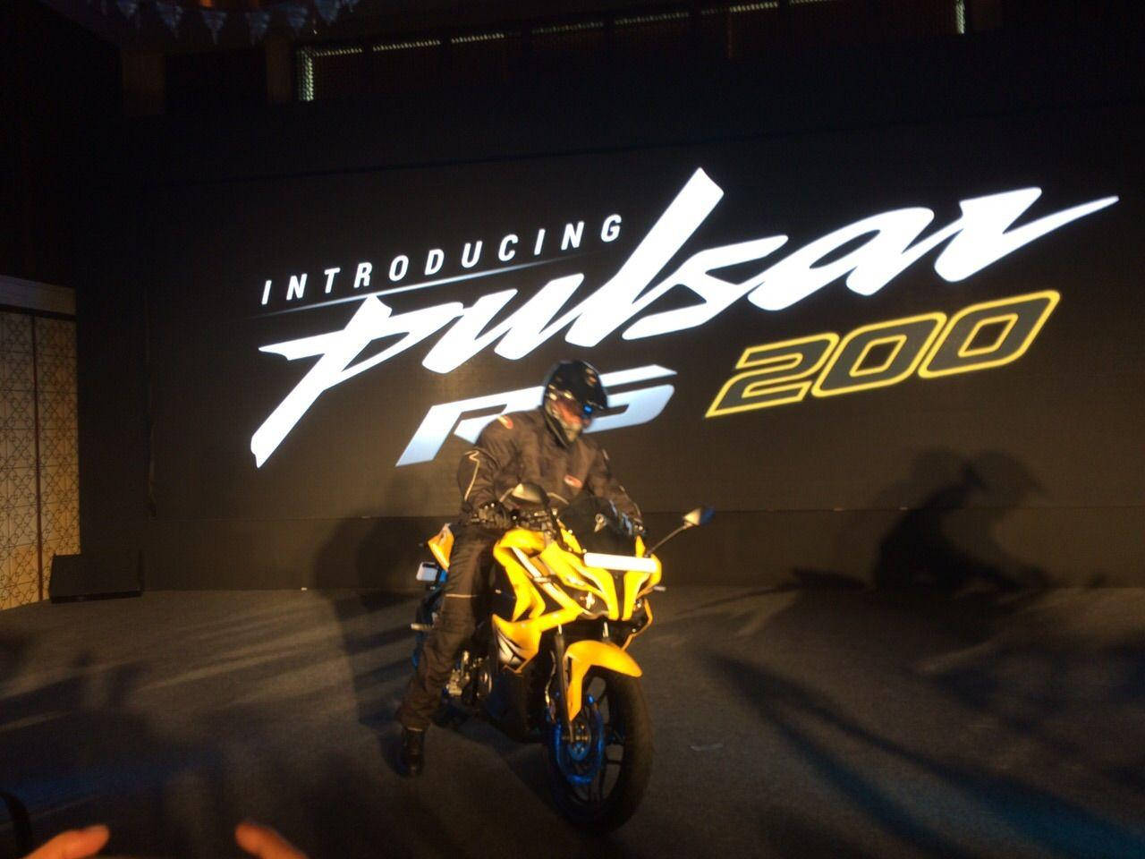 Racing 桌布 壁紙 — 一名全副裝備的騎士在戲劇性的舞台燈光下，坐在一輛充滿活力的黃色Pulsar RS 200摩托車上，姿態優雅。