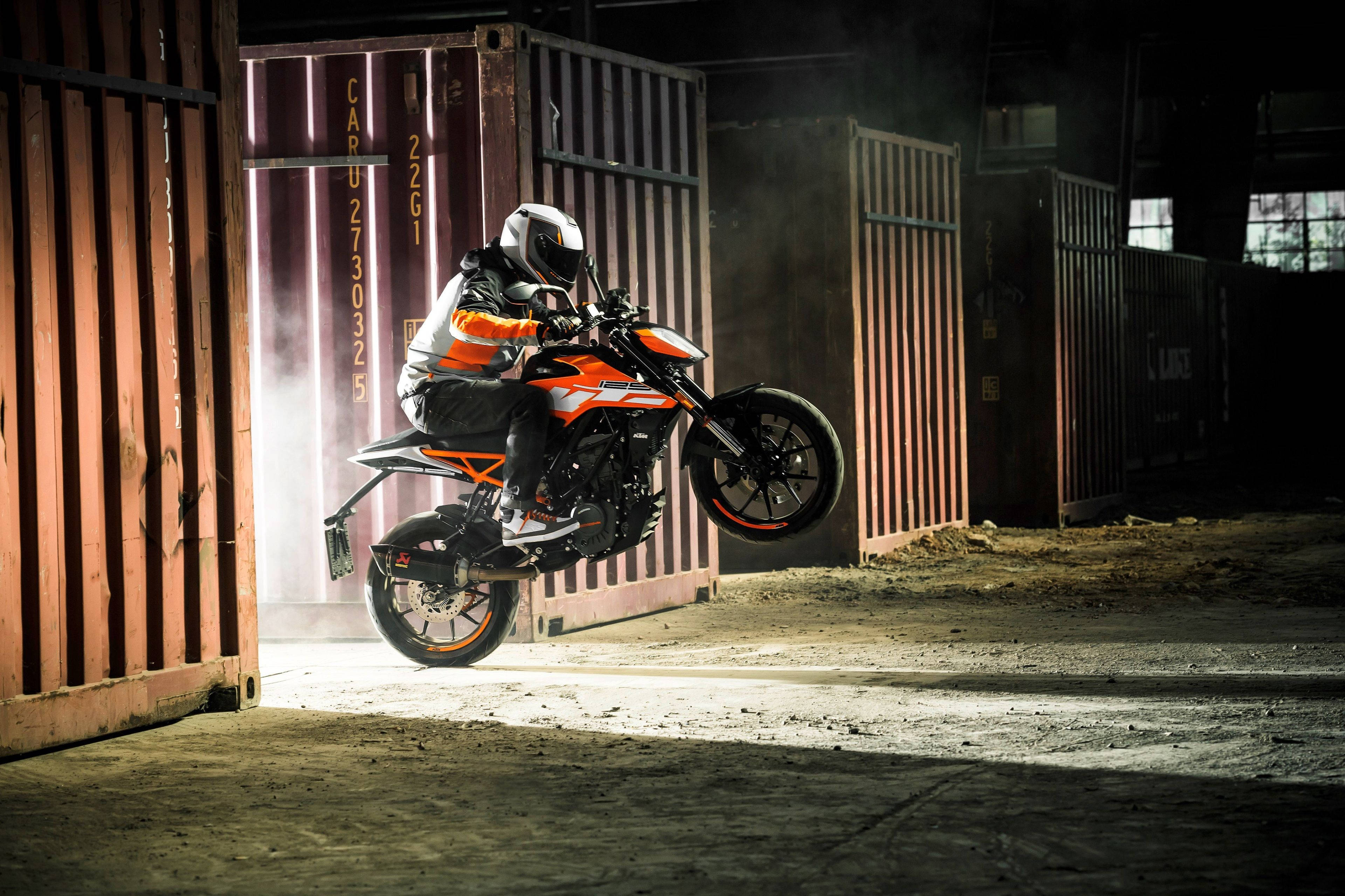 KTM 摩托車 桌布 壁紙 — 一名全副裝備的騎士在昏暗的倉庫中，於生鏽的貨運集裝箱之間，駕駛橙色KTM摩托車表演翹前輪。