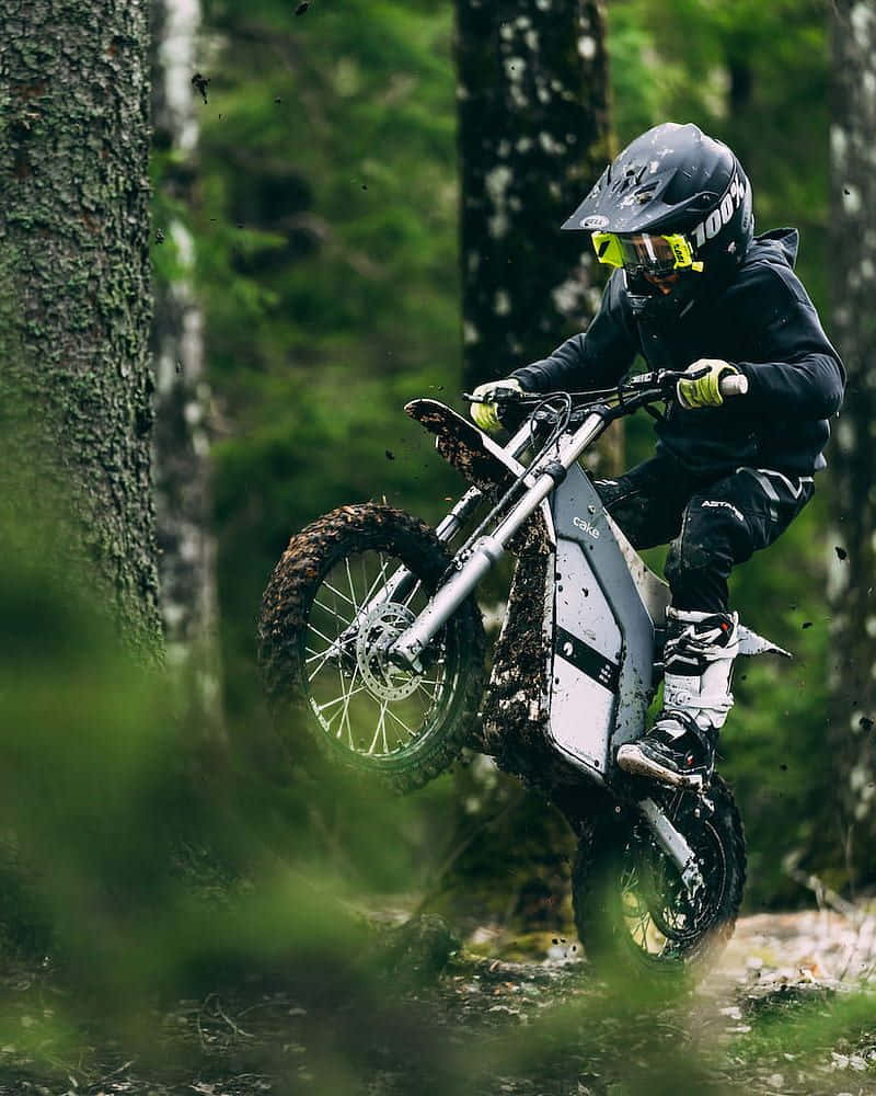 Electric Bike 桌布 壁紙 — 一名全副裝備的騎士在長滿苔蘚的森林中騎著越野摩托車表演翹前輪，泥巴四濺。