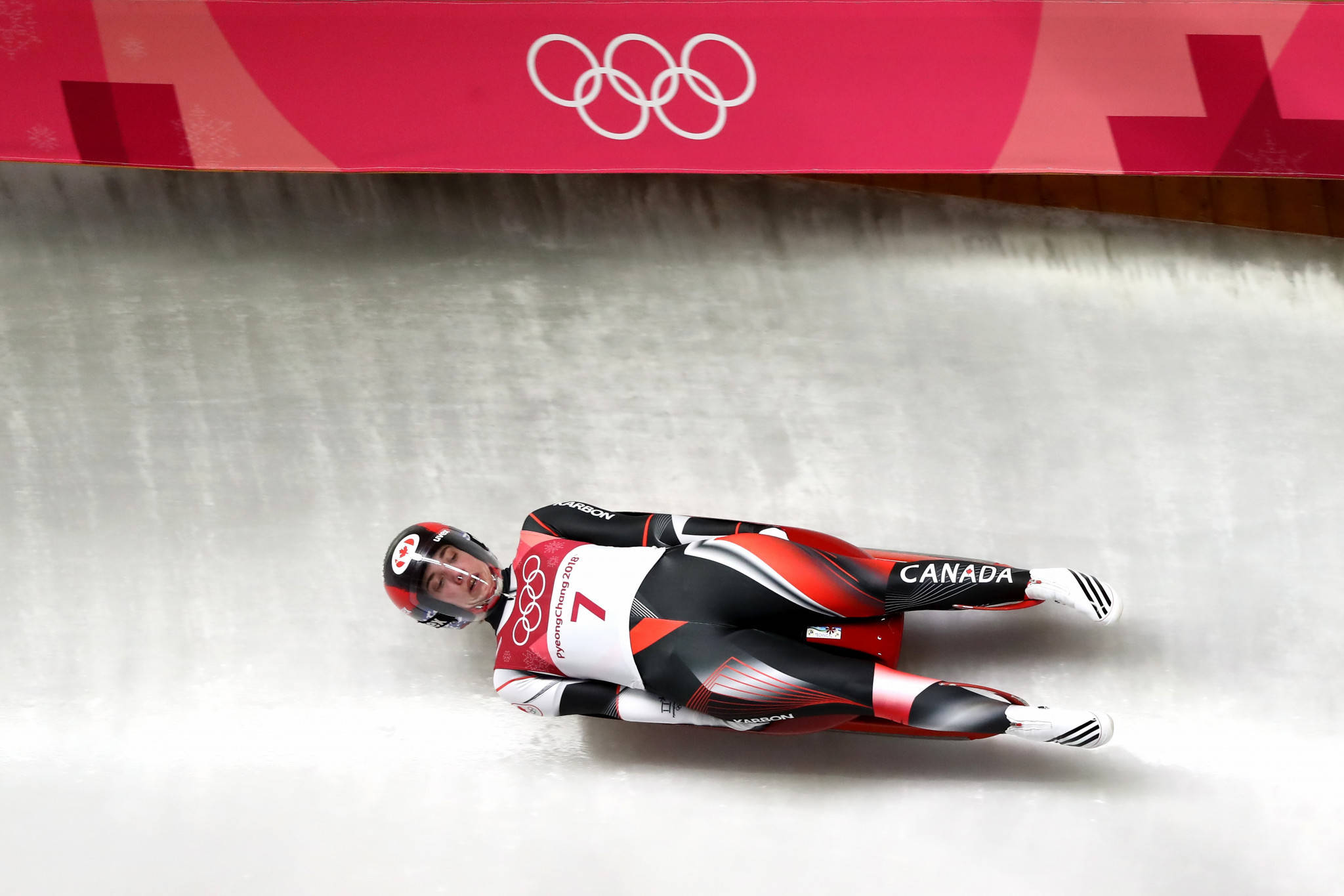 Athletic 桌布 壁紙 — 一名加拿大雪橇運動員身穿紅、黑、白色裝備，在奧運品牌標誌下滑下雪道。