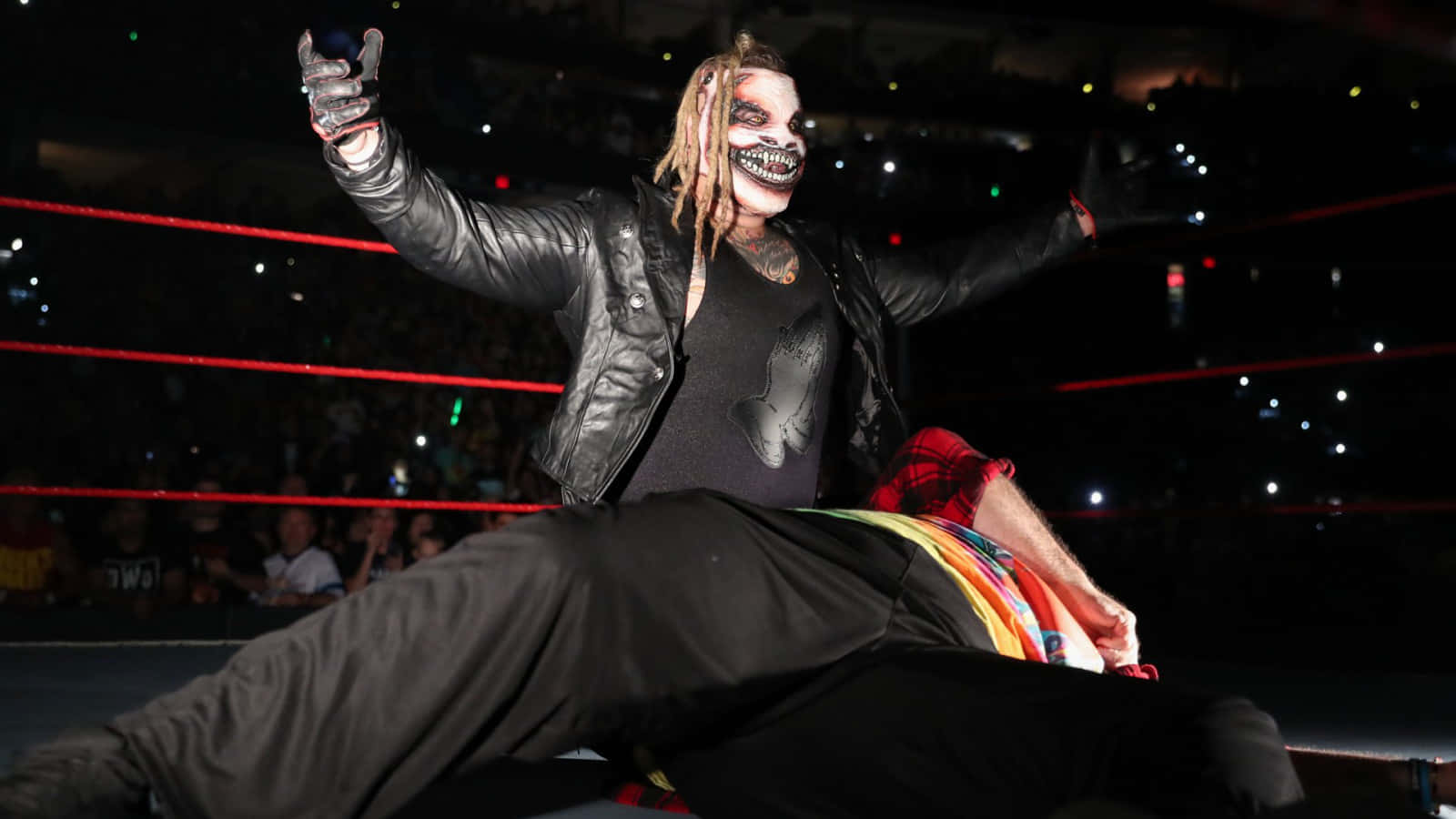 WWE 桌布 壁紙 — 一名化著骷髏妝、留著辮子的摔角手在擂台上高舉雙臂，勝利地站在倒下的對手之上。