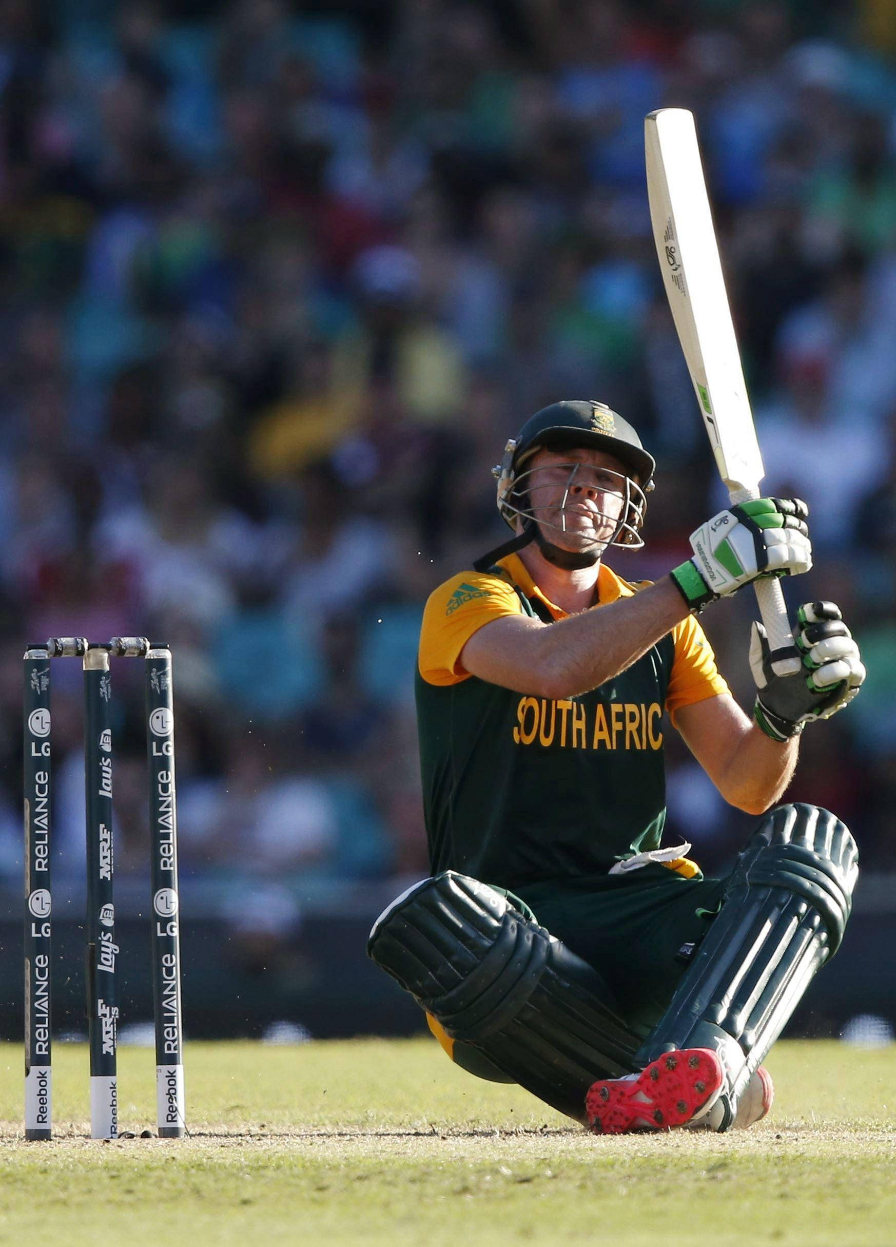 Ab De Villiers 桌布 壁紙 — 一名南非板球擊球手全副裝備跪在草地上，旁邊是三柱門，高舉球棒，在一個坐滿觀眾的體育場內。