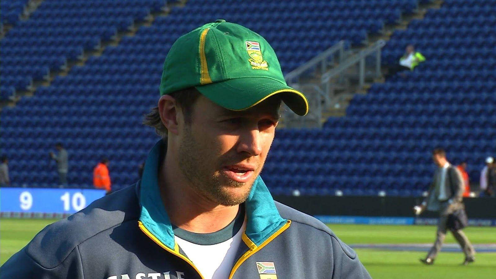 Ab De Villiers 桌布 壁紙 — 一名南非板球運動員戴著綠色帽子、穿著藍色外套，站在陽光普照的體育場草地上，身後是空無一人的藍色座位。