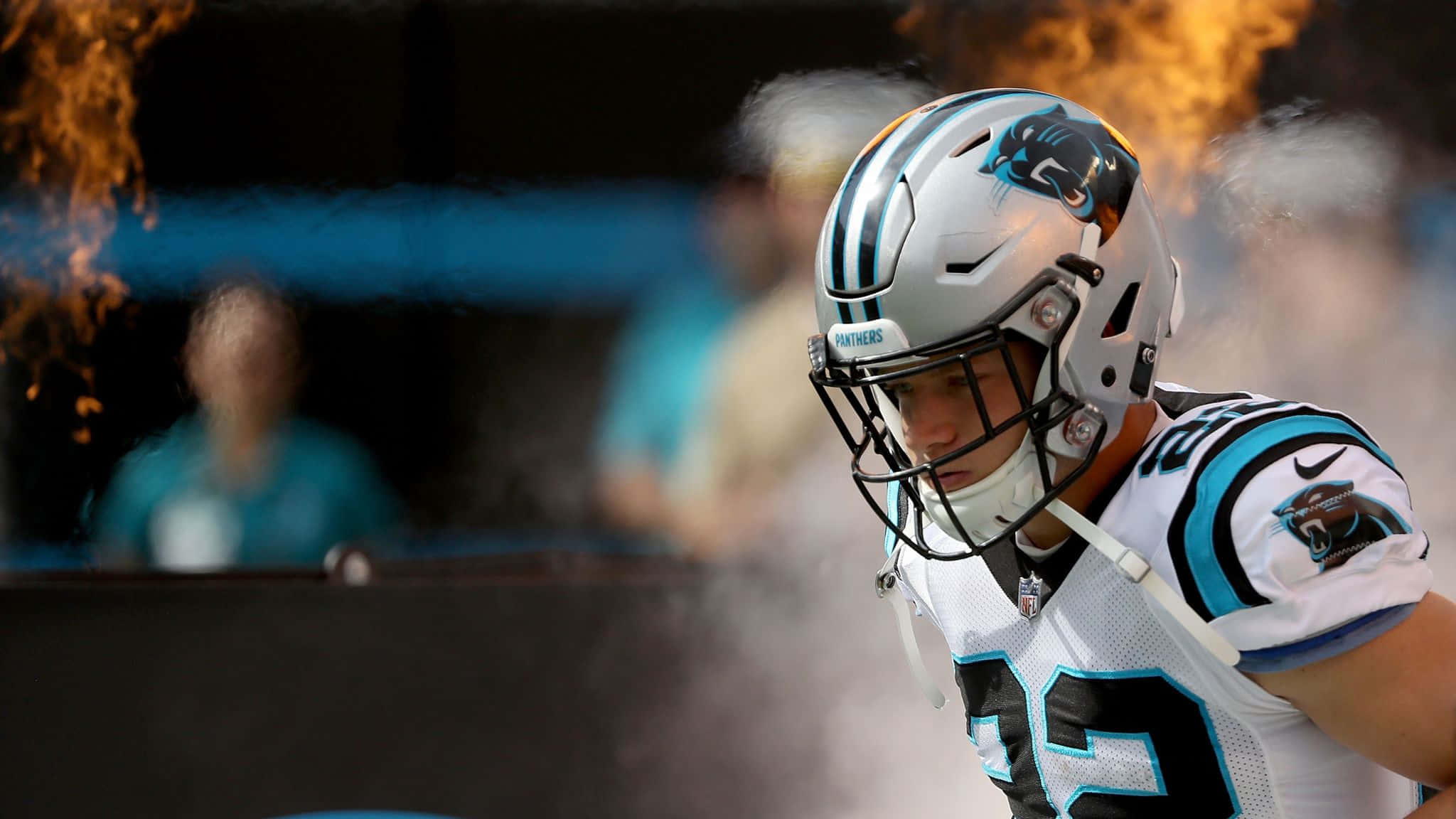 Christian Mccaffrey 桌布 壁紙 — 一名卡羅萊納黑豹隊美式足球員身穿白色和青綠色球衣，從煙霧中出現，背景有火焰。