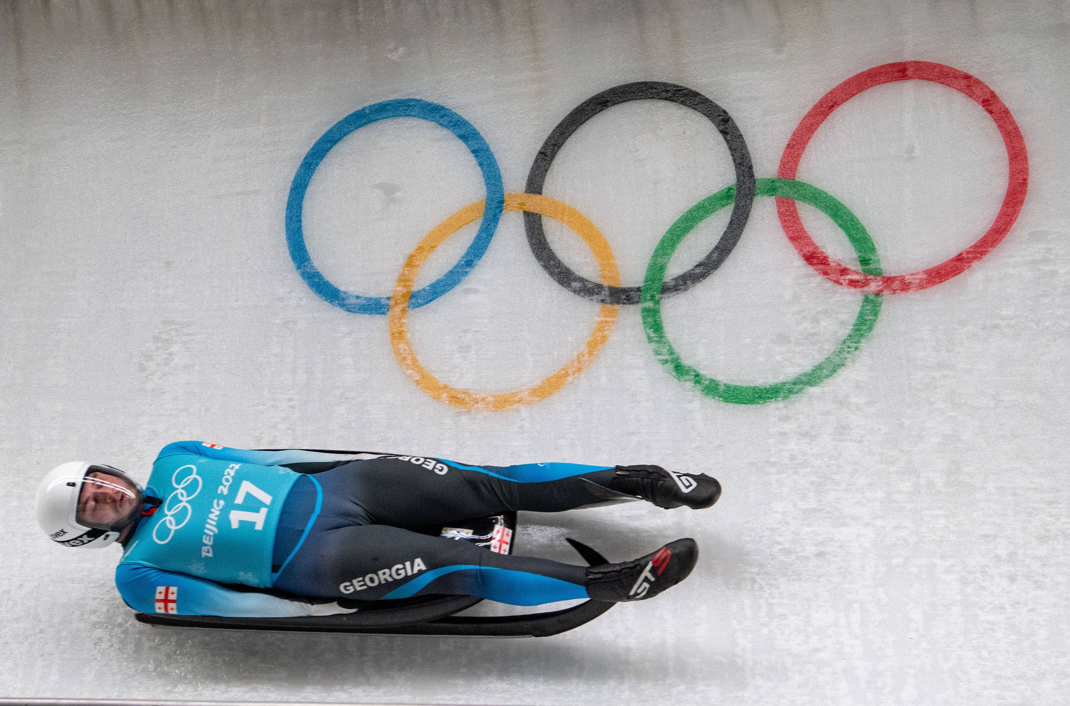 Athletic 桌布 壁紙 — 一名喬治亞雪橇運動員身穿藍黑色裝備，在背後牆上繪有奧運五環的冰道上飛馳。
