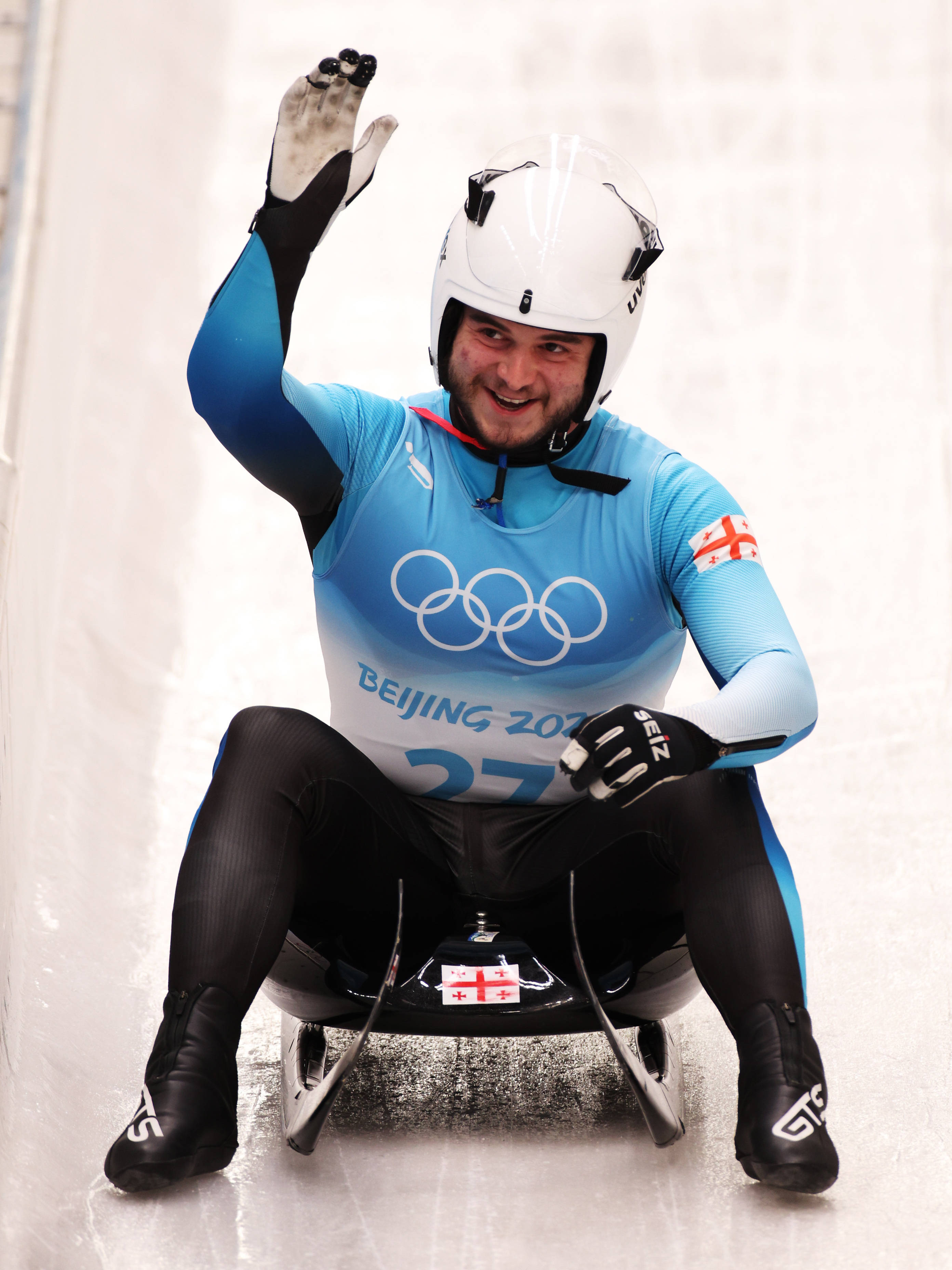 Athletic 桌布 壁紙 — 一名喬治亞雪車選手身穿藍色奧運制服，在北京2022年冬奧會期間在冰道上從雪車上愉快地揮手。