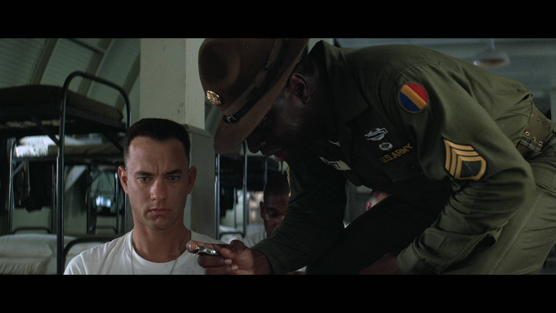 forrest 桌布 壁紙 — 一名嚴肅的士兵身穿橄欖色制服，在軍營房間內俯身於一名坐著的男子，該男子手持剃刀刀片。