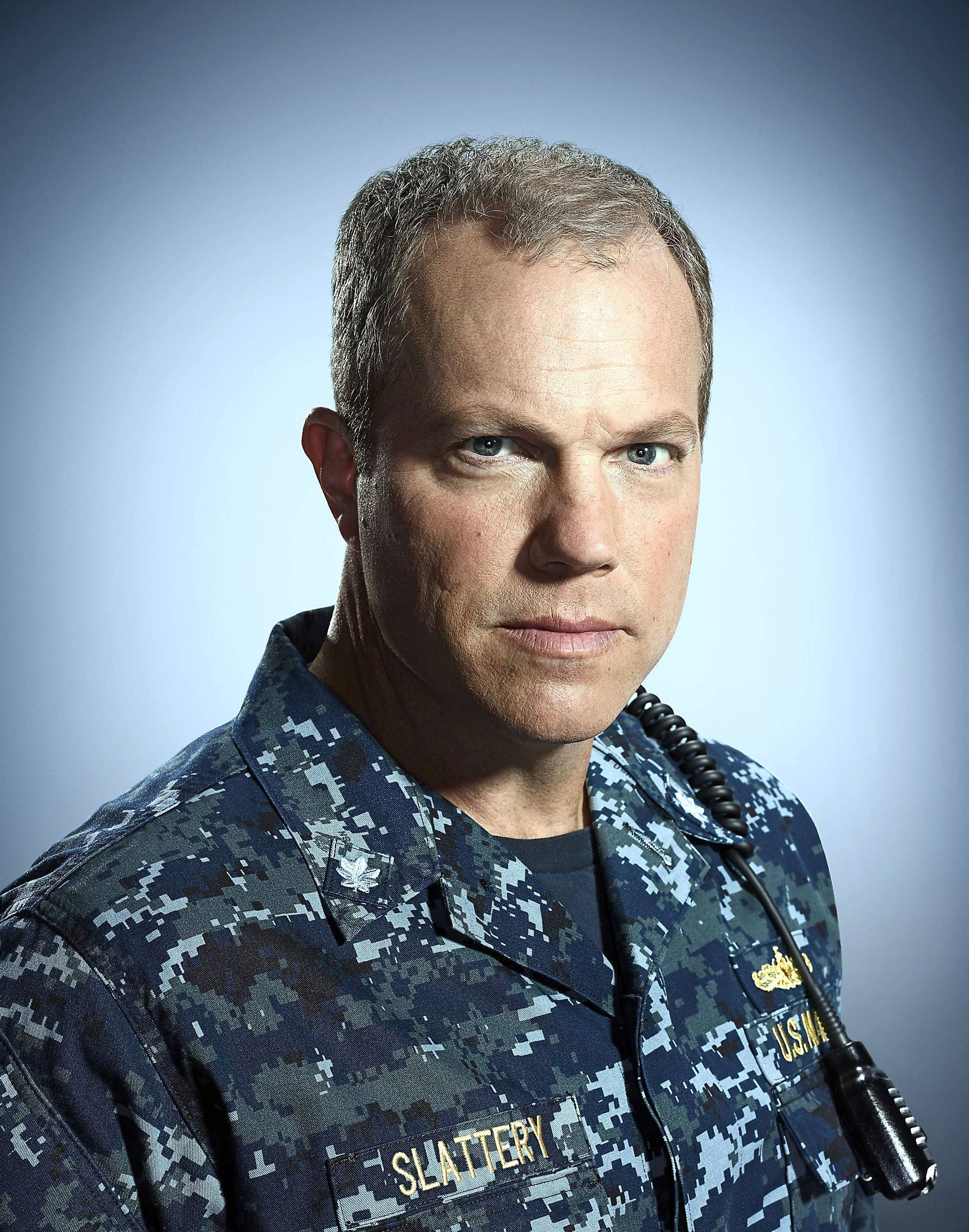 adam baldwin 桌布 壁紙 — 一名嚴肅的男子身穿海軍藍數位迷彩制服，佩戴姓名牌和無線電耳機，背景為漸層藍色。