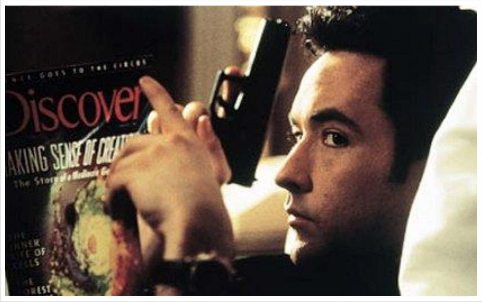 John Cusack 桌布 壁紙 — 一名在床上的男人在溫暖燈光下，專注地將槍指向一本《Discover》雜誌封面。