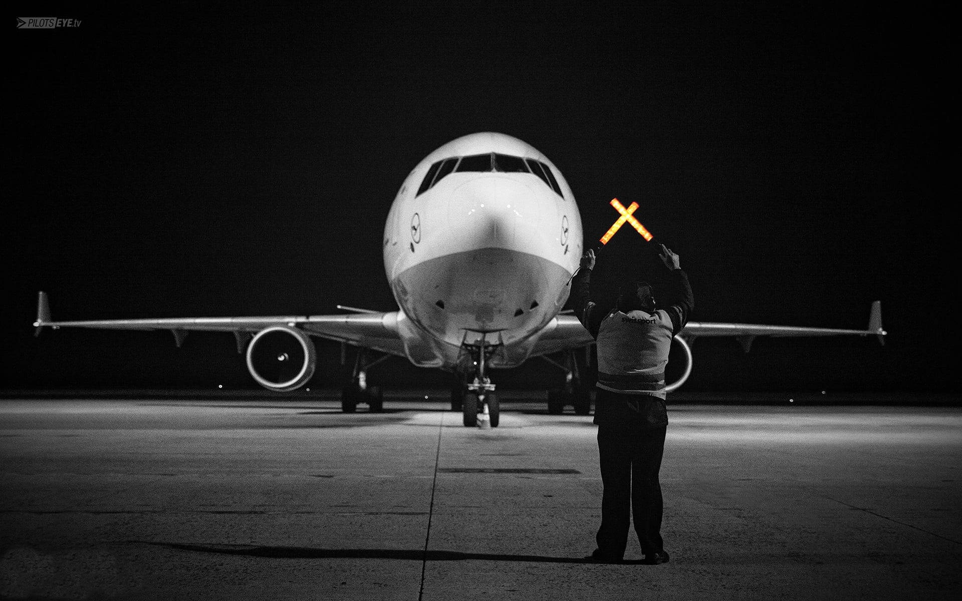 飛機 桌布 壁紙 — 一名地勤人員在夜間黑暗的停機坪上，在一架大型飛機前手持一個發光的橙色X形燈光。