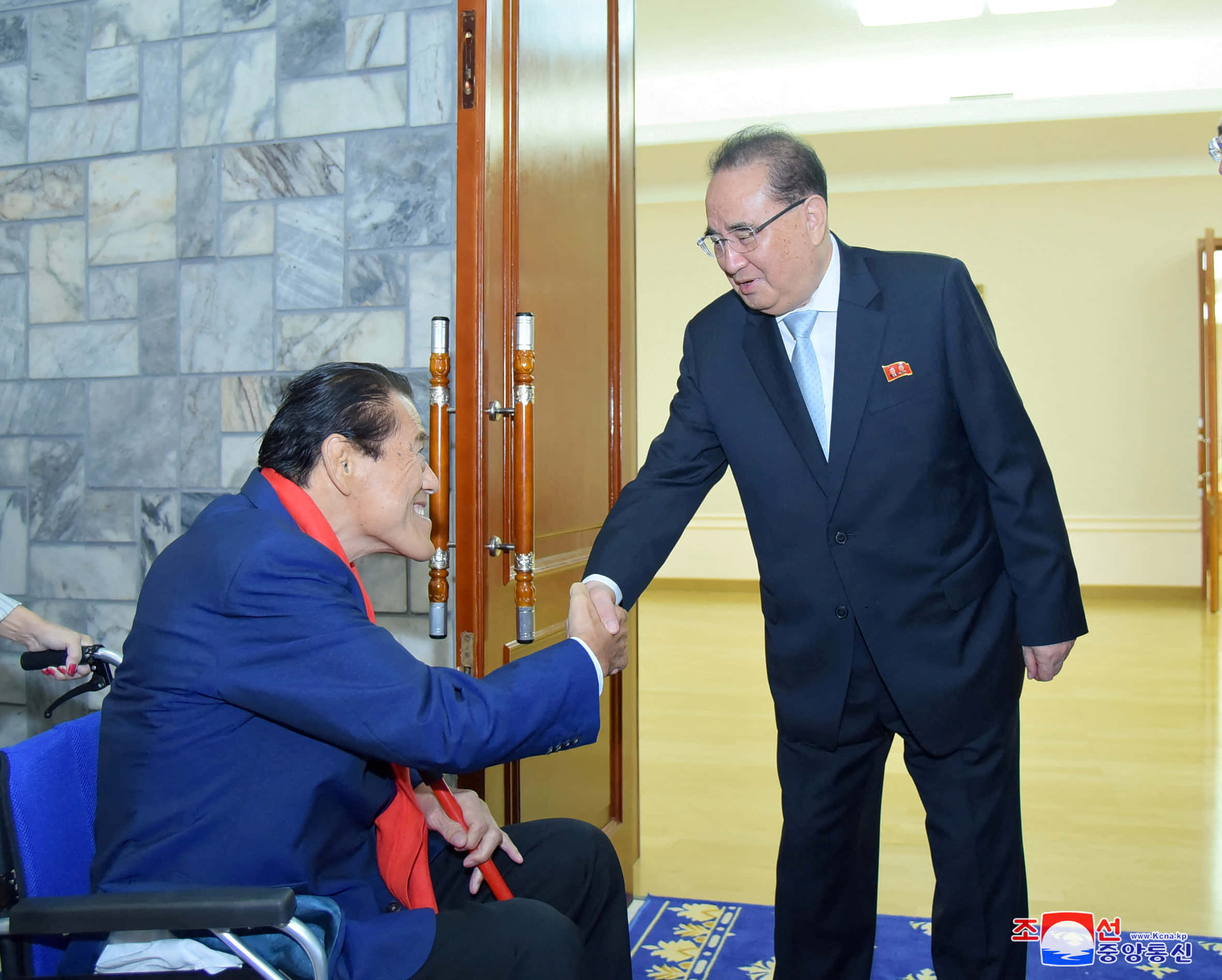運動 桌布 壁紙 — 一名坐輪椅的男子與一名身穿西裝站立的男子在一個有大理石牆壁和木門的正式房間內握手。