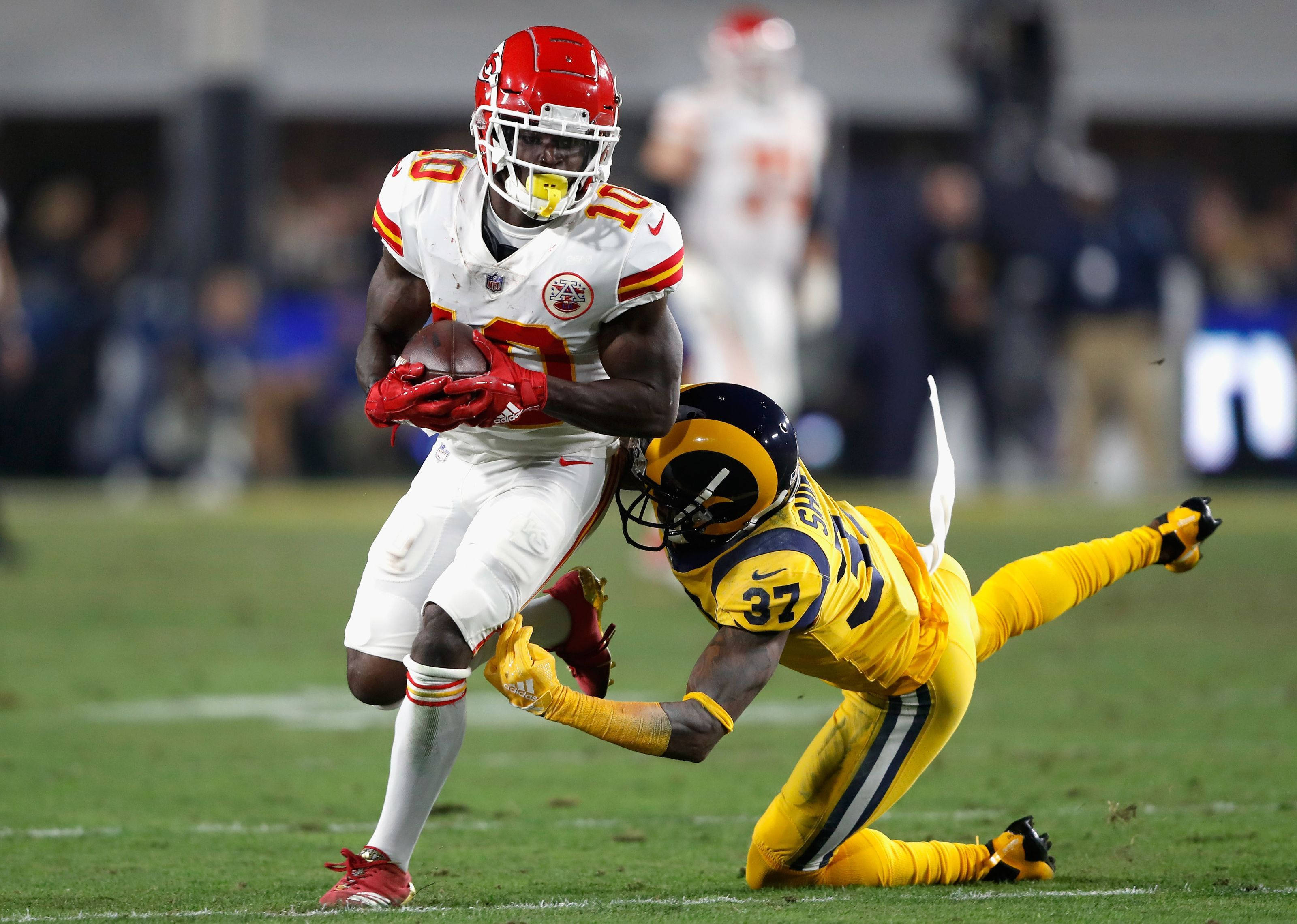 Tyreek Hill 桌布 壁紙 — 一名堪薩斯城酋長隊的紅白球員帶著美式足球奔跑，而洛杉磯公羊隊的黃色防守球員在綠色場地上飛撲擒抱他。