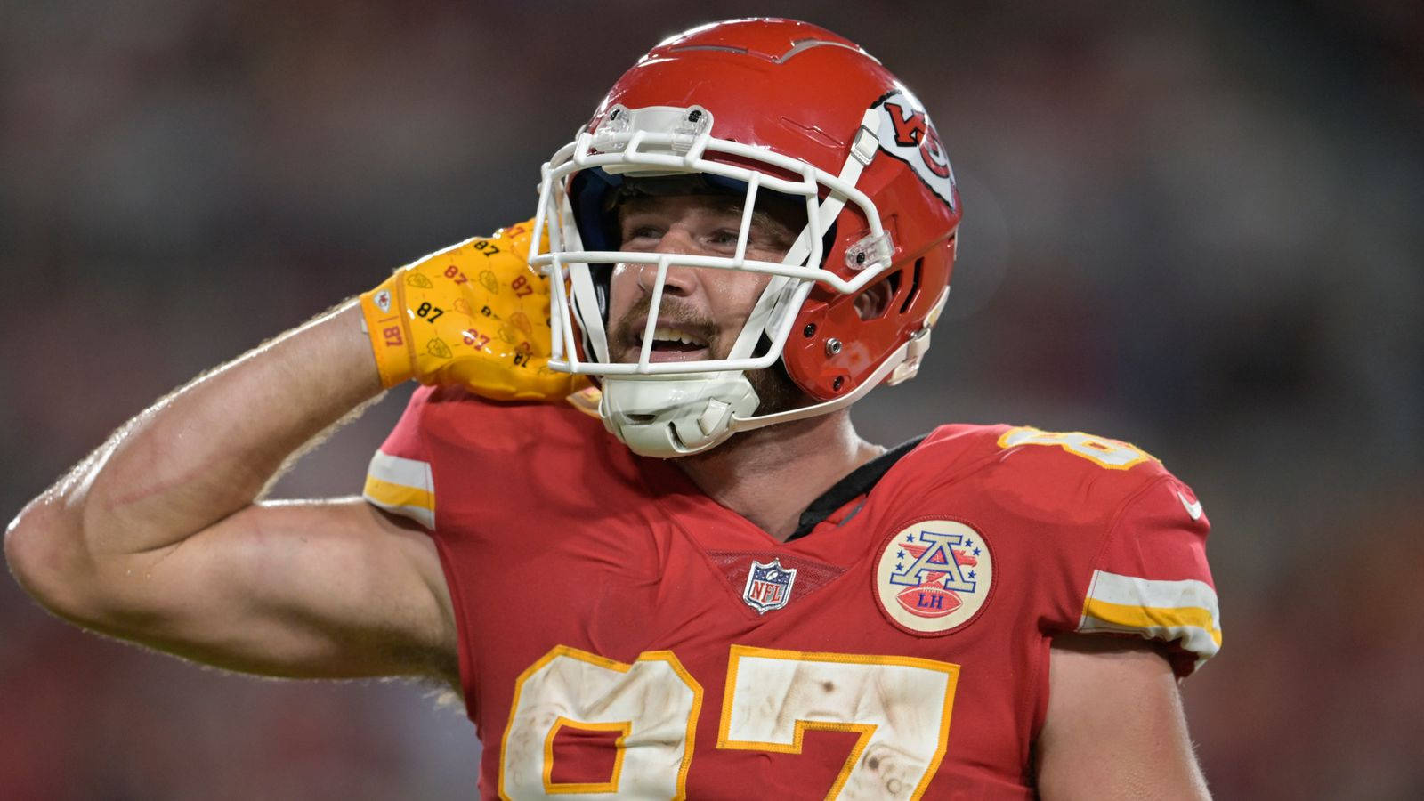 Travis Kelce 桌布 壁紙 — 一名堪薩斯城酋長隊的美式足球員身穿紅色球衣和頭盔，在明亮燈光下的體育場內將戴著手套的手舉到頭部。