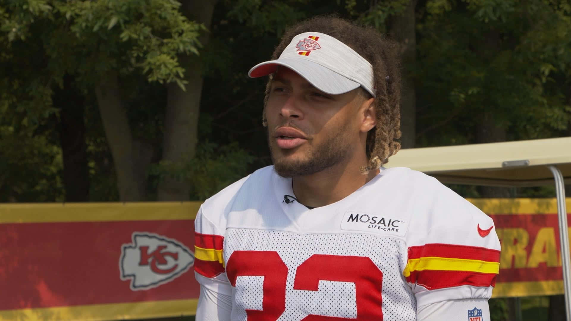 Tyrann Mathieu 桌布 壁紙 — 一名堪薩斯城酋長隊的美式足球員身穿帶有紅色和黃色點綴的白色球衣，站在戶外，身後有樹木和帶有品牌標誌的屏障。