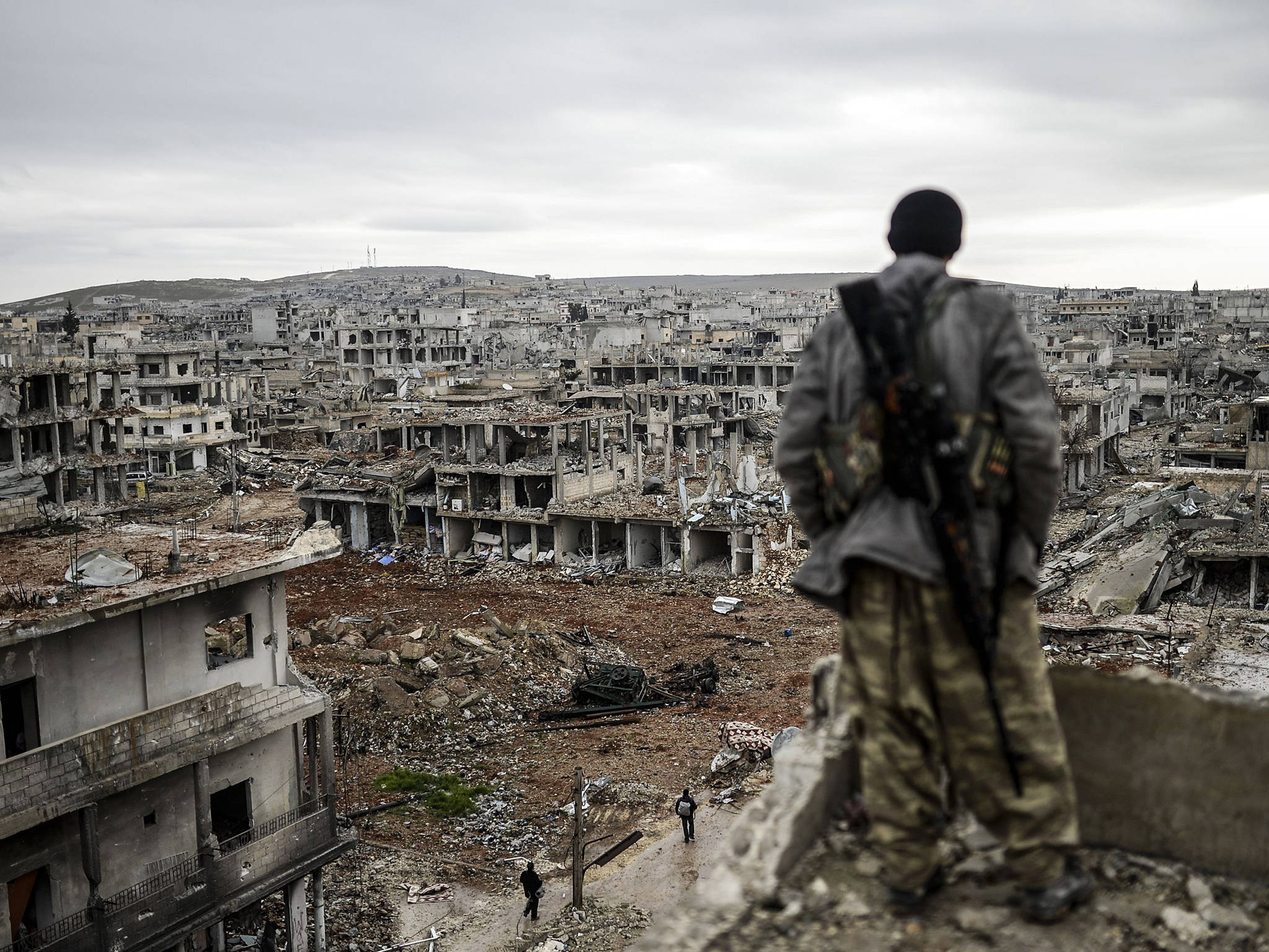 Syria 桌布 壁紙 — 一名士兵站在瓦礫上，俯瞰灰色天空下廣闊的廢墟城市景觀。