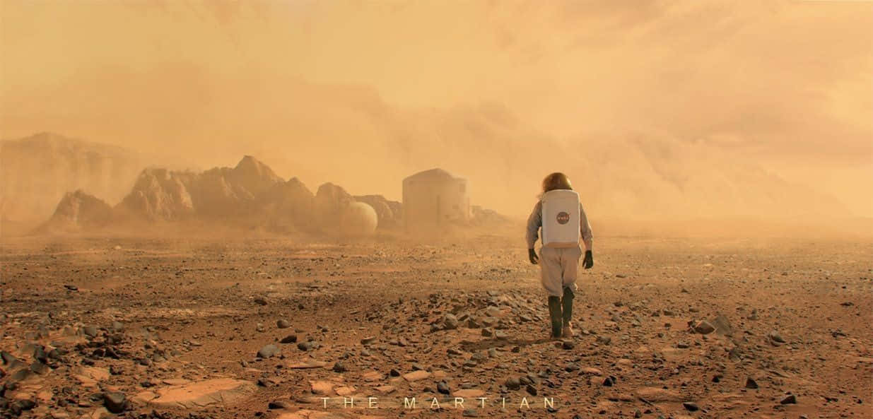 The Martian 桌布 壁紙 — 一名太空人在塵土飛揚的火星景觀中獨自走向遠處的棲息地，天空是朦朧的橙色。