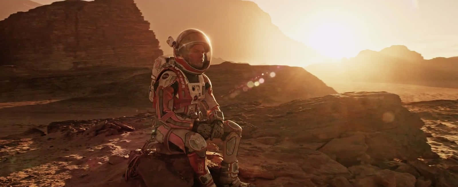The Martian 桌布 壁紙 — 一名太空人獨自坐在岩石密布的外星行星上，金色的太陽在遠山後升起，投下長長的影子。
