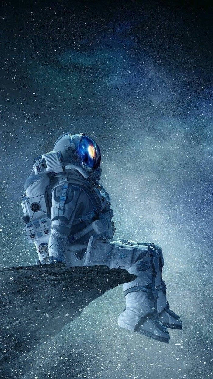 酷炫星系 桌布 壁紙 — 一名太空人獨自坐在岩石露頭上，背景是繁星點點的宇宙，星雲在深藍和紫色中發光。
