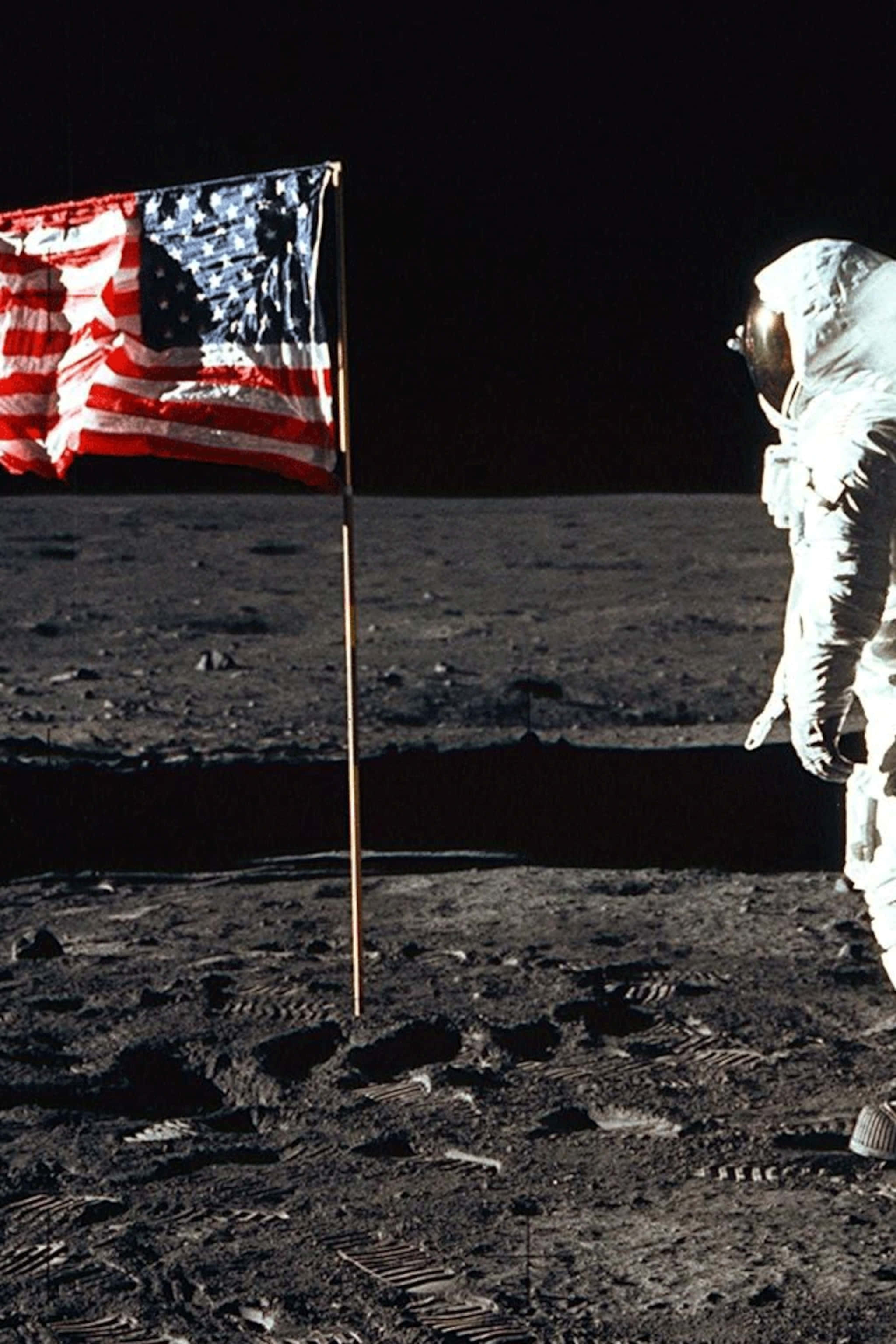 Moon Landing 桌布 壁紙 — 一名太空人站在月球灰色表面上飄揚的美國國旗旁，背景是黑色天空。
