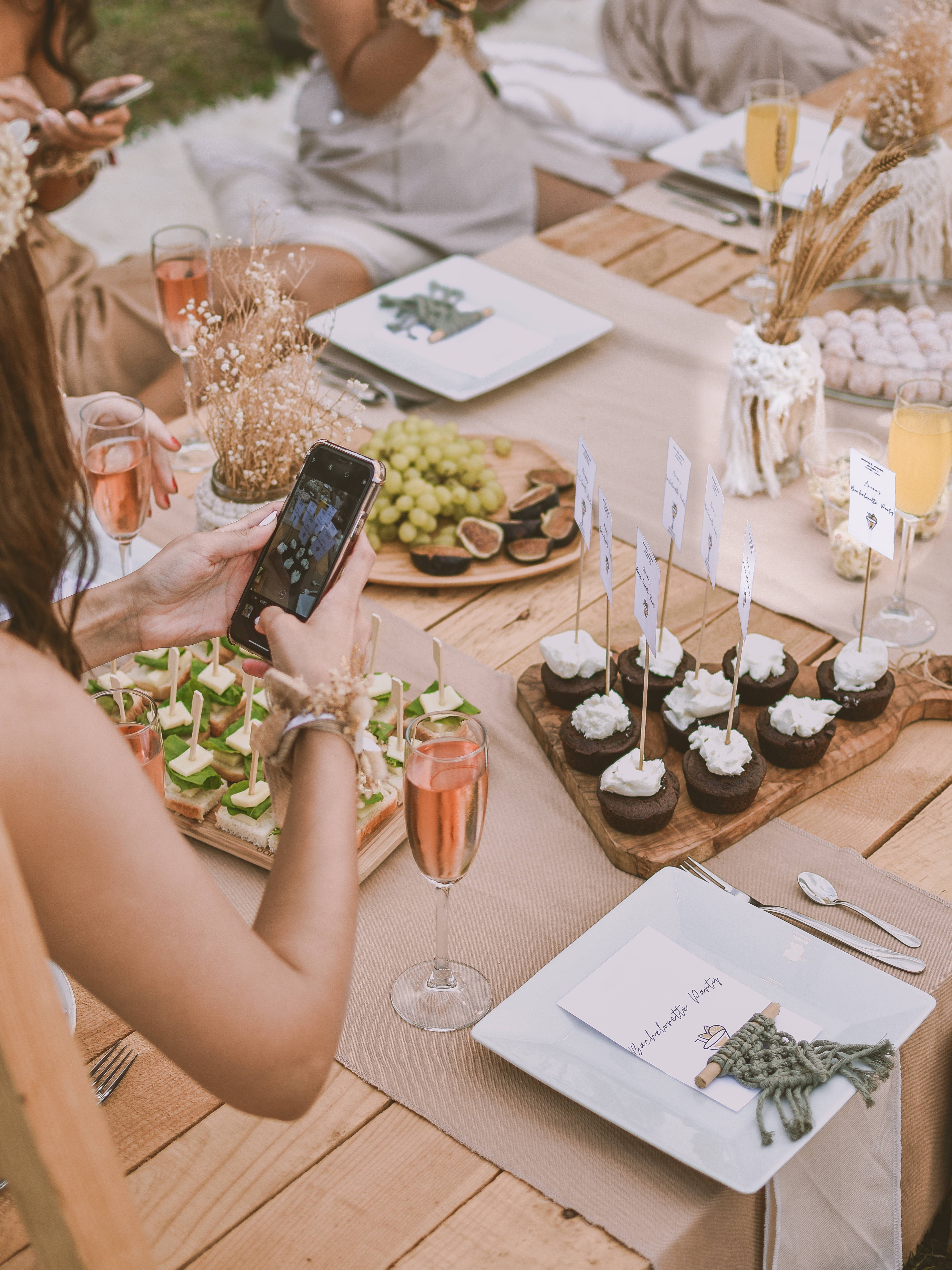Bachelorette 桌布 壁紙 — 一名女士正在拍攝一張擺滿開胃菜和玫瑰酒的鄉村風格戶外野餐桌照片。