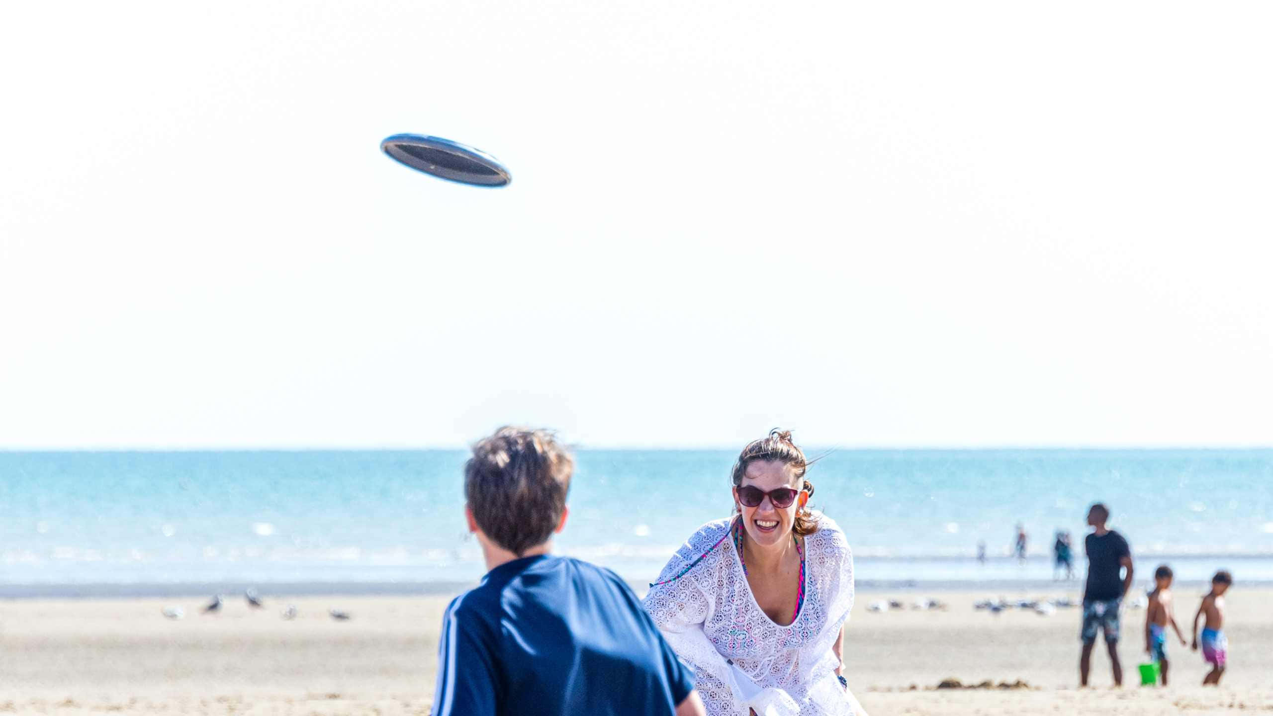 Frisbee 桌布 壁紙 — 一名女士笑著準備接住飛行的飛盤，背景是陽光燦爛的海灘、碧綠的海洋和零星的海灘遊客。