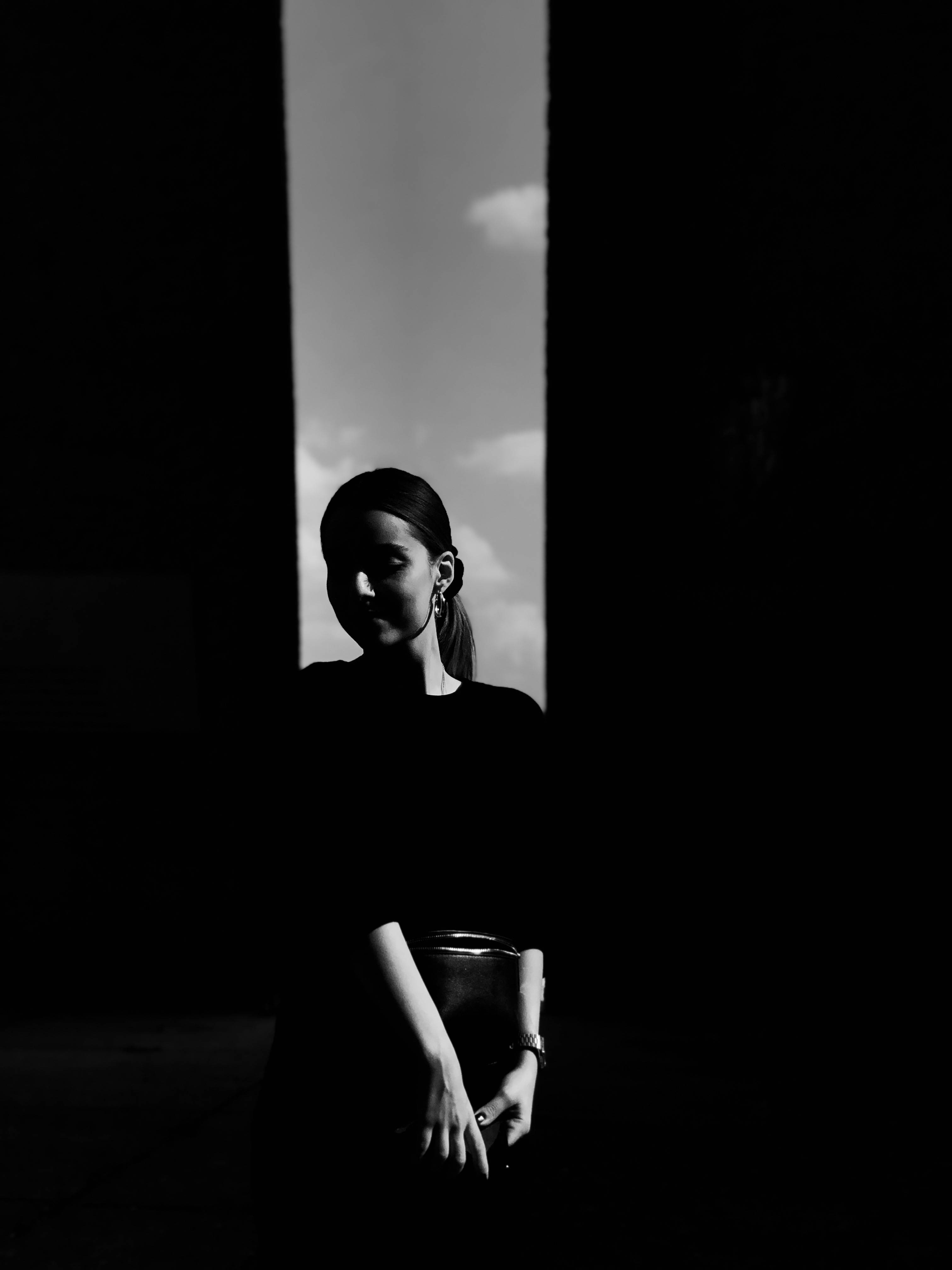 Black And Grey iPhone 桌布 壁紙 — 一名女子以剪影姿態站在狹窄的垂直窗框內，背景是陰雲密佈的天空，呈現高對比度的黑白效果。
