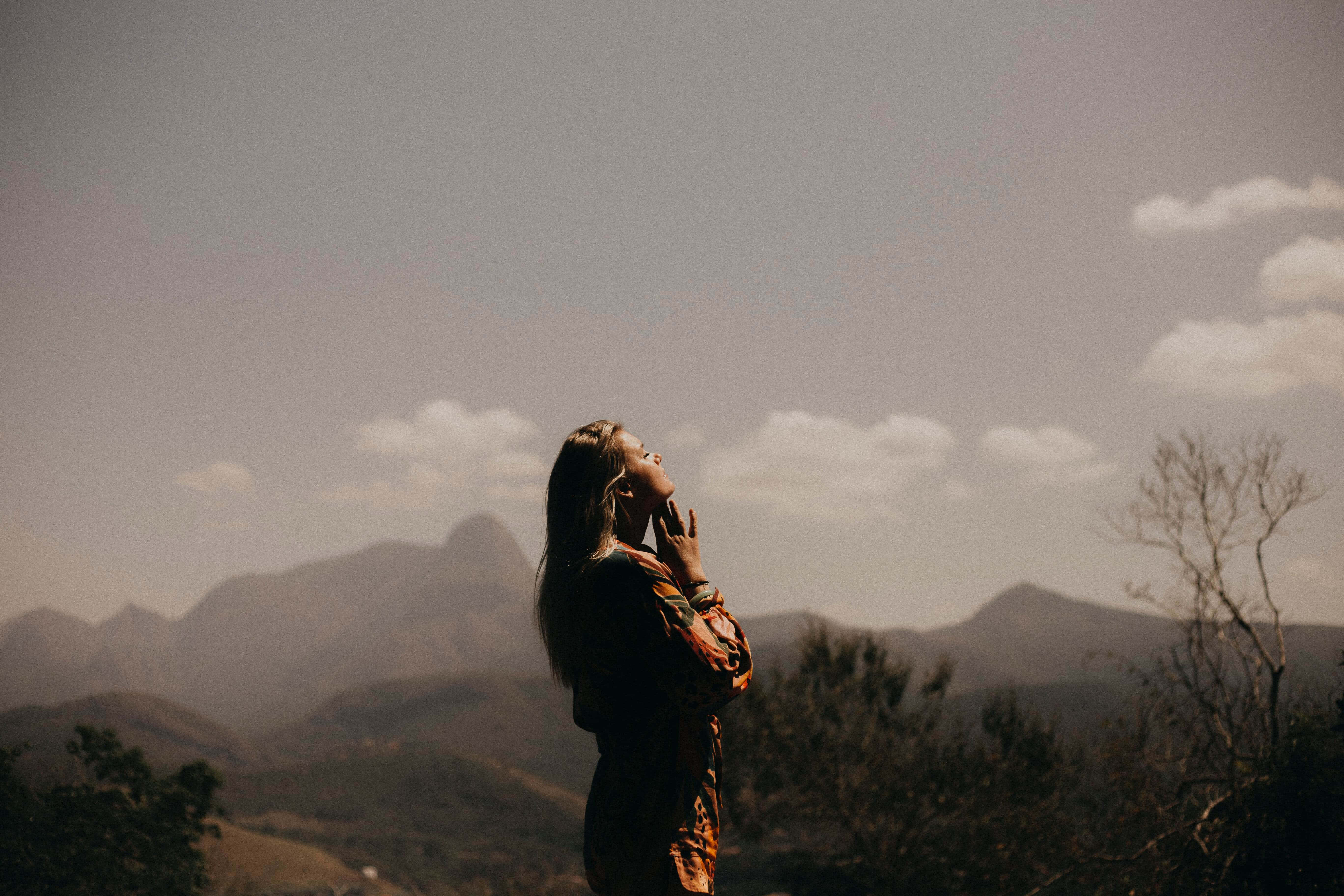 Calming 桌布 壁紙 — 一名女子以剪影姿態站在遠山前，雙手靠近臉龐，仰望柔和的多雲天空。
