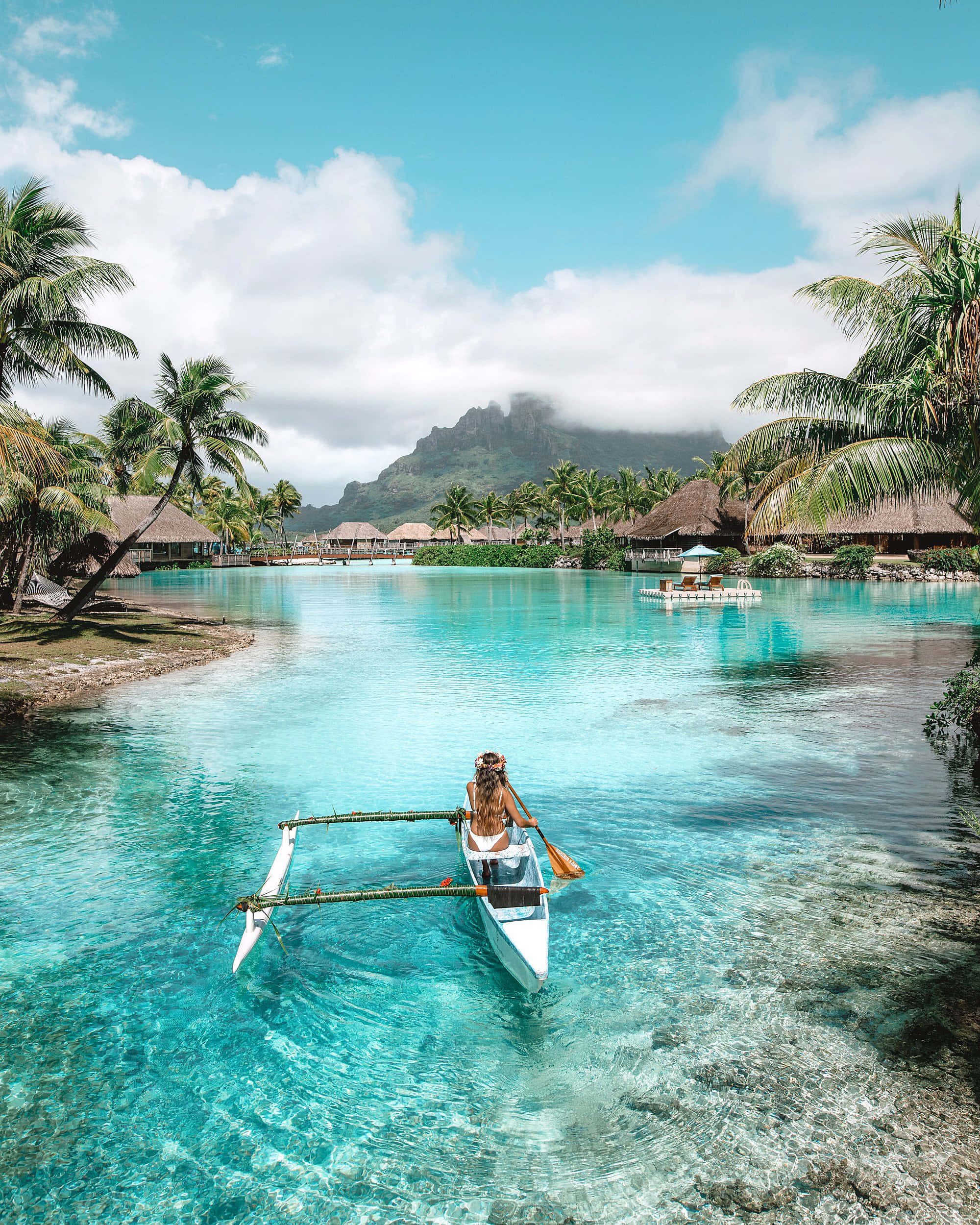 Bora Bora Beach 桌布 壁紙 — 一名女子划著傳統的舷外支架獨木舟，穿過清澈碧綠的海水，周圍環繞著棕櫚樹和水上平房，頭頂是明亮的藍天。