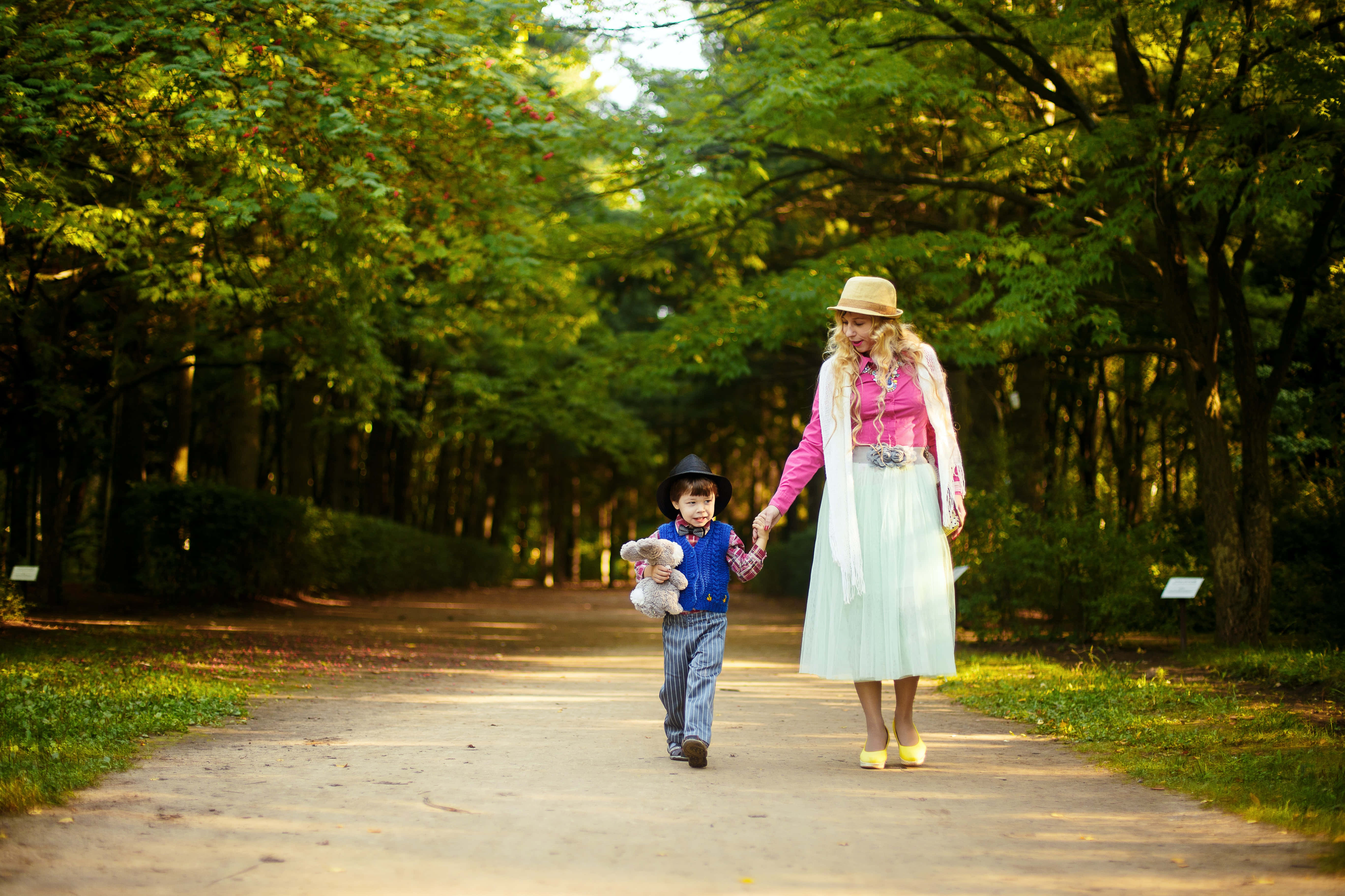 Mom 桌布 壁紙 — 一名女子和一個孩子手牽手，走在綠意盎然的公園裡一條陽光普照、綠樹成蔭的小徑上。