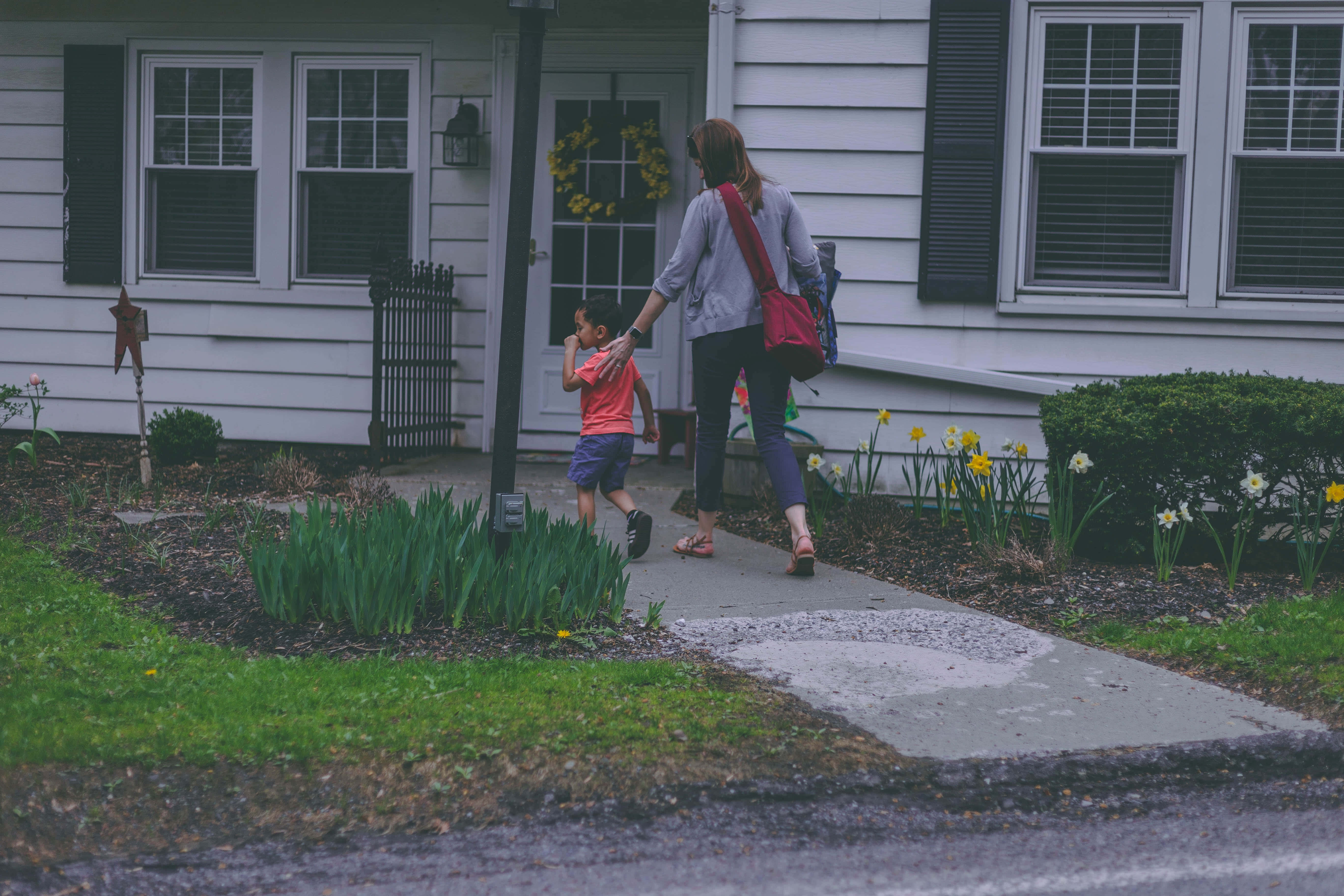 Mom 桌布 壁紙 — 一名女子和一個孩子手牽手走在人行道上，經過一間白色房子，院子裡有水仙花盛開。