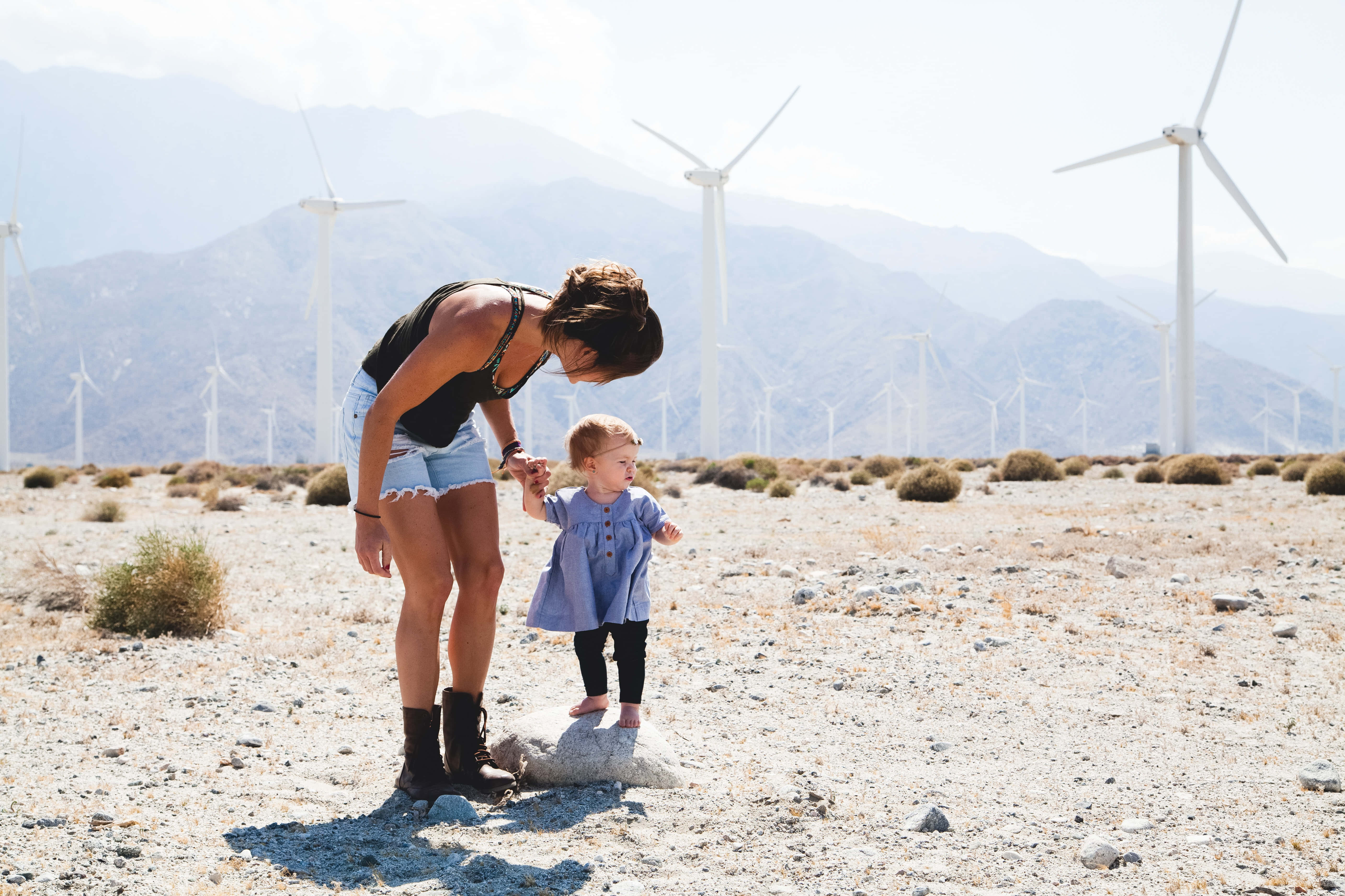Mom 桌布 壁紙 — 一名女子和一個幼兒在陽光明媚的沙漠中手牽手，背景有風力渦輪機和山脈。