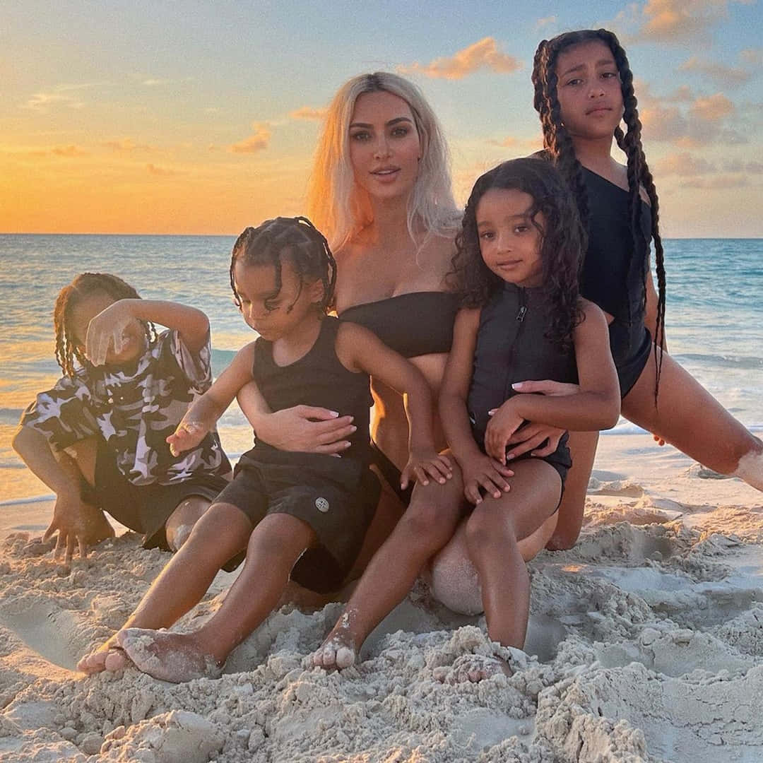 Beach Family 桌布 壁紙 — 一名女子和她的四個孩子一起坐在沙灘上，夕陽西下，溫暖的金色光芒映照在海面上。