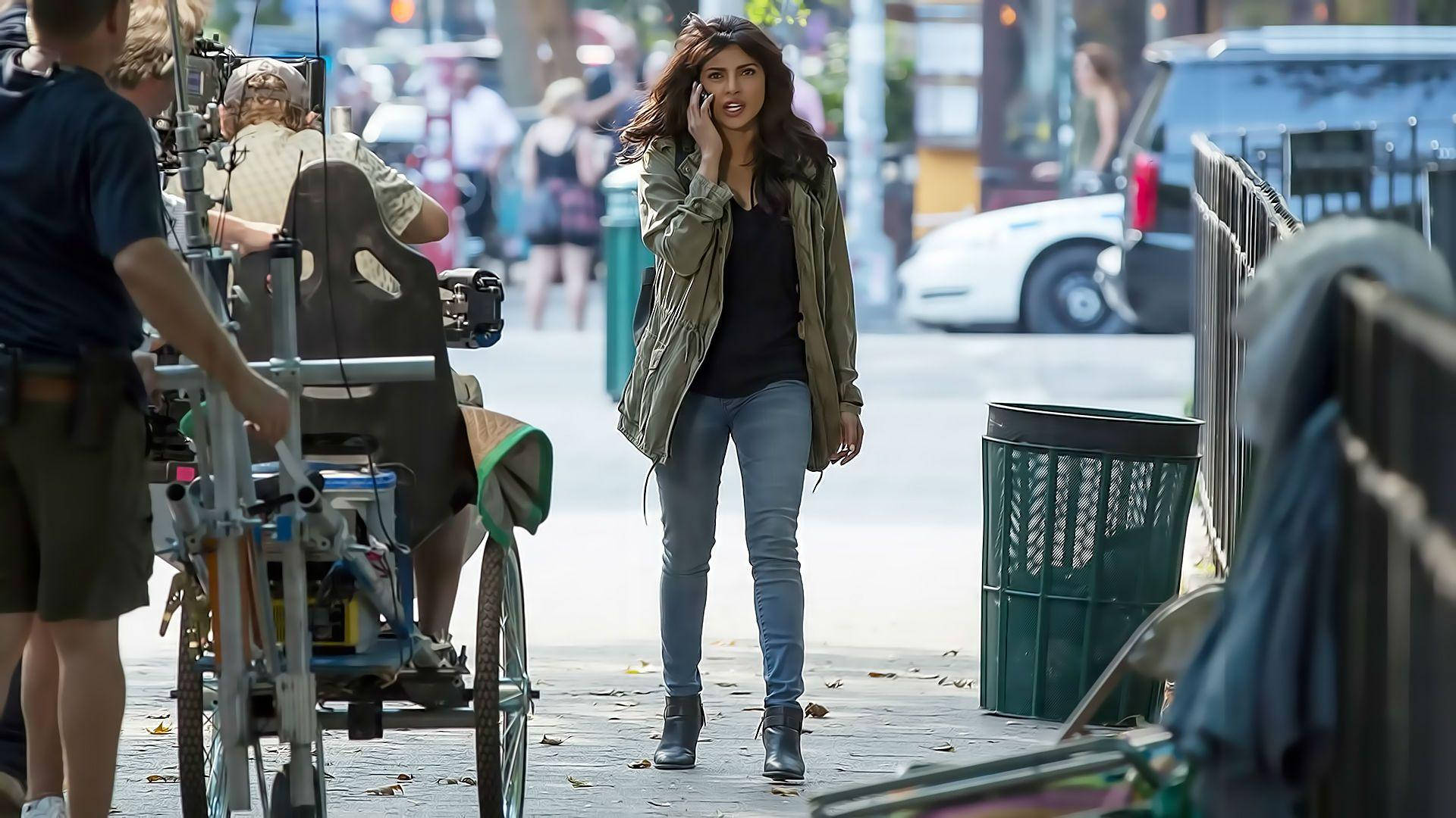 Priyanka 桌布 壁紙 — 一名女子在城市人行道上行走，一邊講電話，附近有電影工作人員操作設備。