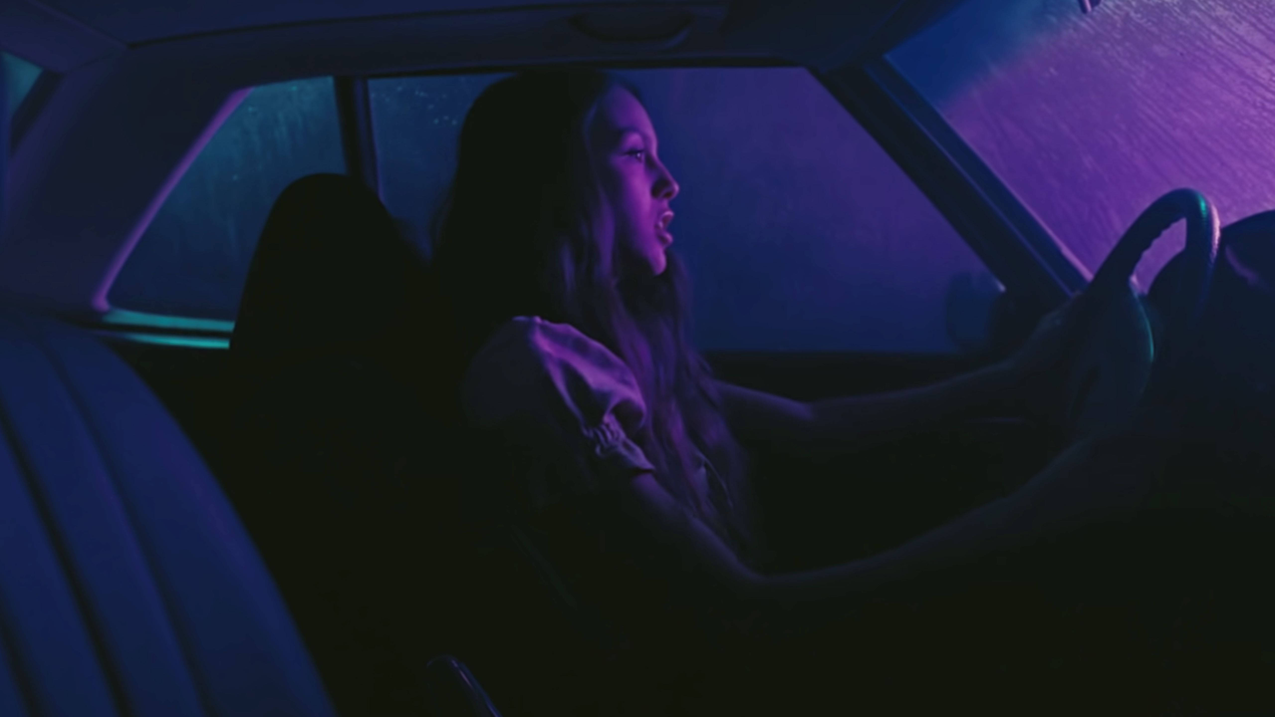 奧莉維亞·羅德里戈 桌布 壁紙 — 一名女子在夜間駕車穿過雨中的霓虹紫色和藍色街燈，車內視角。