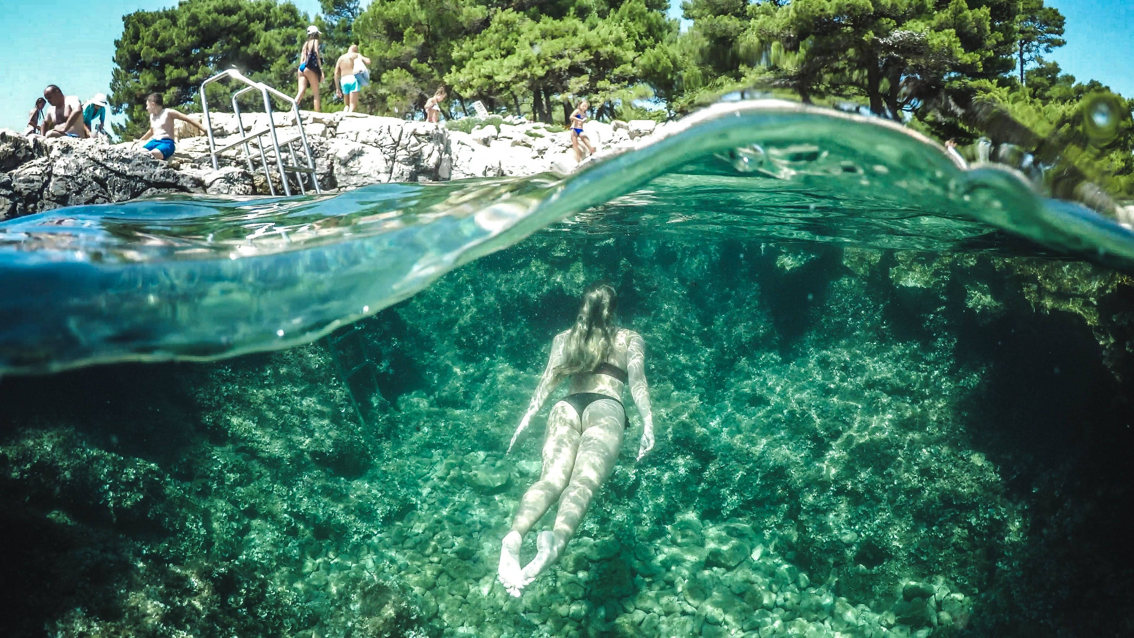 Snorkeling 桌布 壁紙 — 一名女子在水晶般清澈的碧綠海中游泳，而其他人則在上方陽光普照的岩石海岸上放鬆。