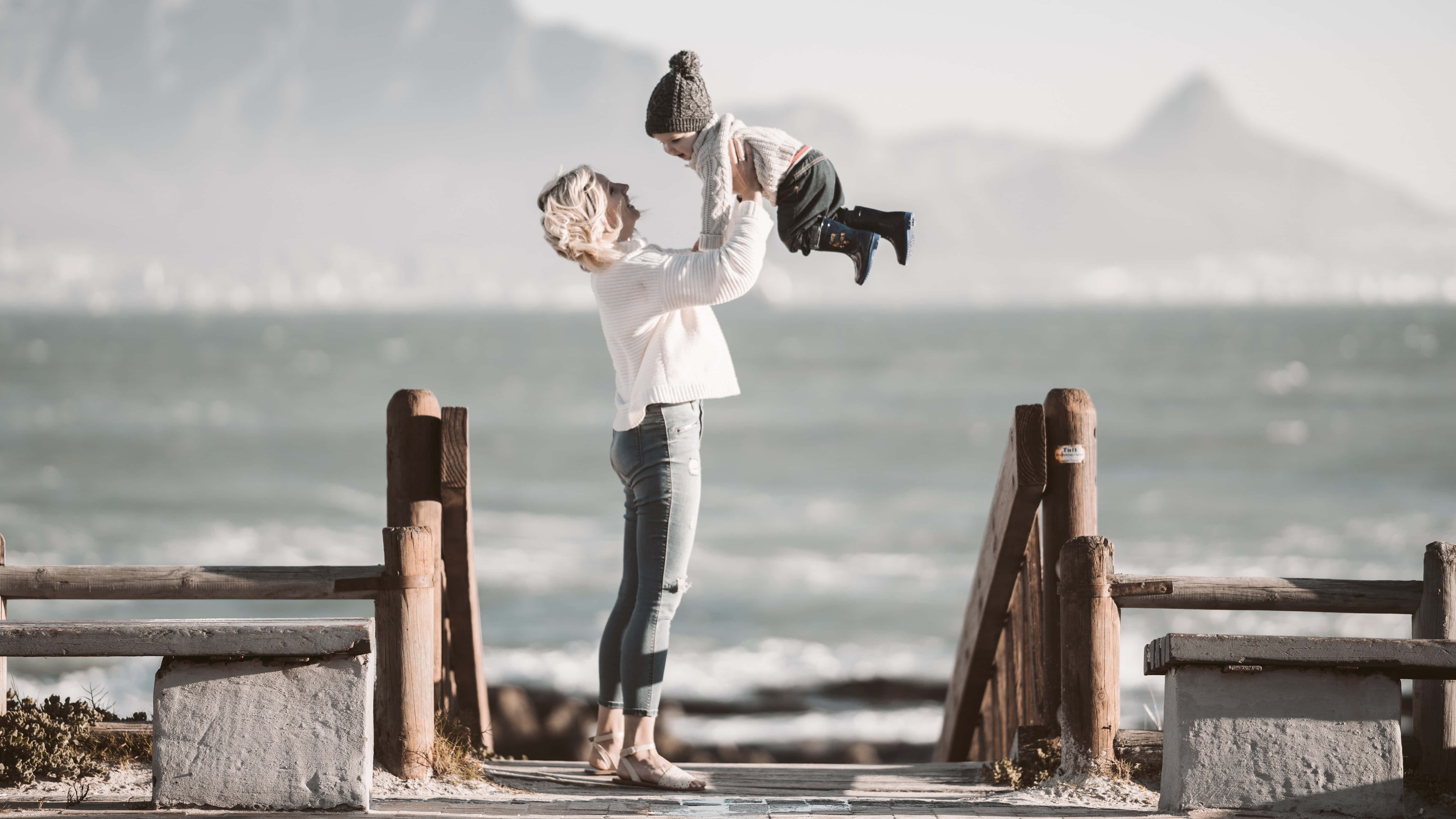 Mom 桌布 壁紙 — 一名女子在海洋波浪和遠山的背景下，高興地舉起一個戴著針織帽的幼兒。