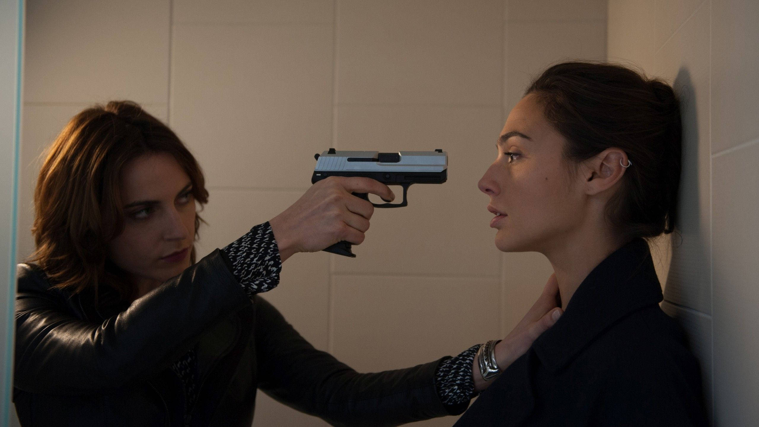 Antje Traue 桌布 壁紙 — 一名女子在瓷磚牆前持槍指向另一名女子的頭部，形成緊張對峙。