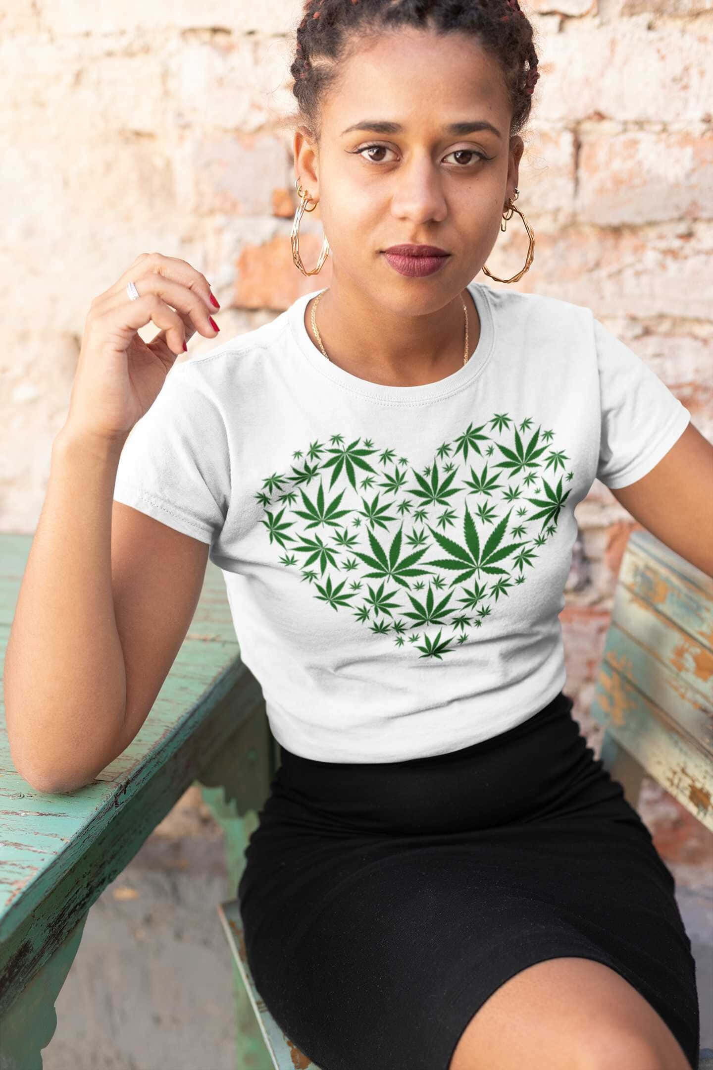 大麻 桌布 壁紙 — 一名女子坐在一張風化的綠色長椅上，身穿白色T恤，上面有綠色大麻葉心形圖案，背景是磚牆。