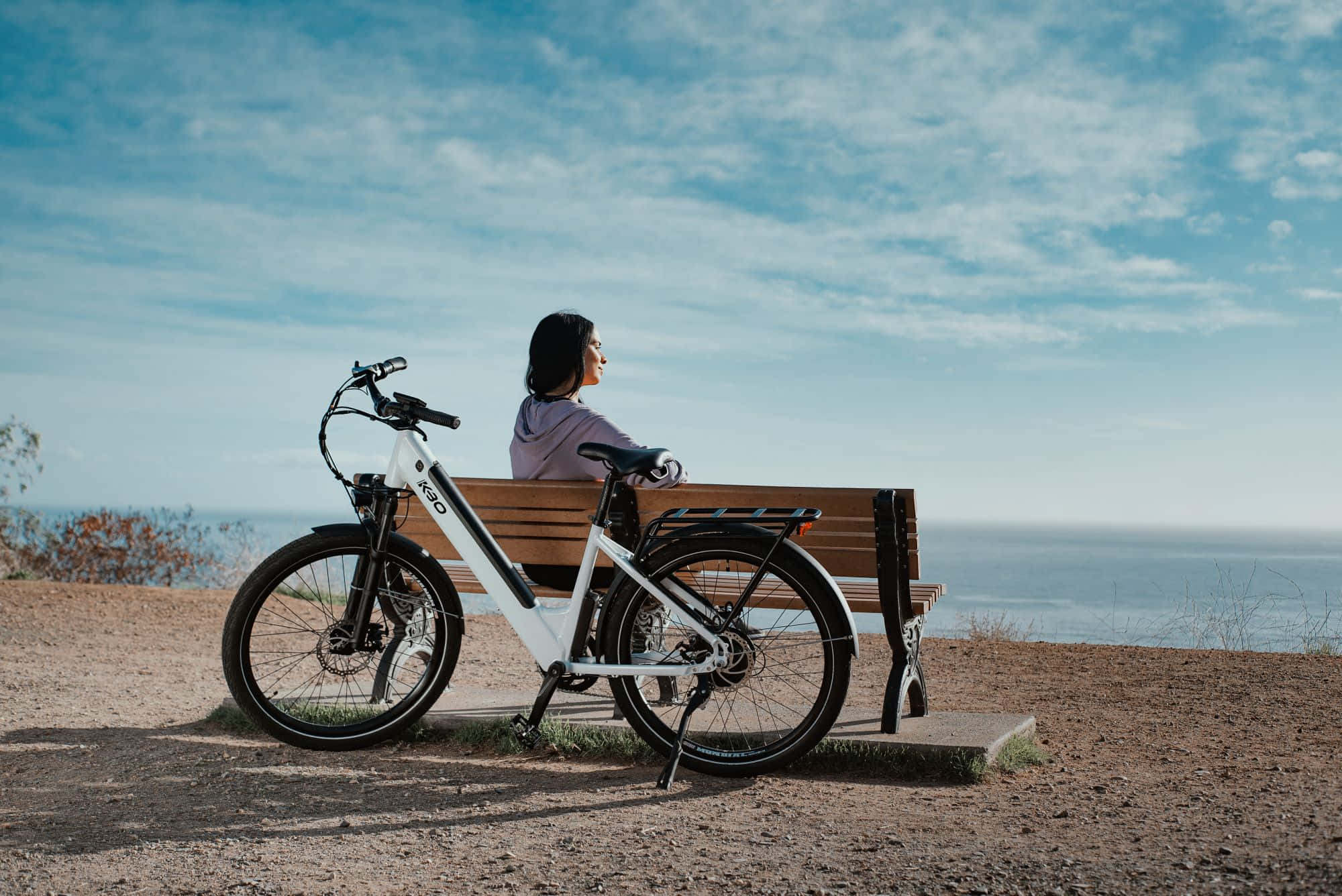 Electric Bike 桌布 壁紙 — 一名女子坐在可俯瞰海洋的木長椅上，一輛白色電動自行車停在她旁邊，頭頂是明亮的藍天。