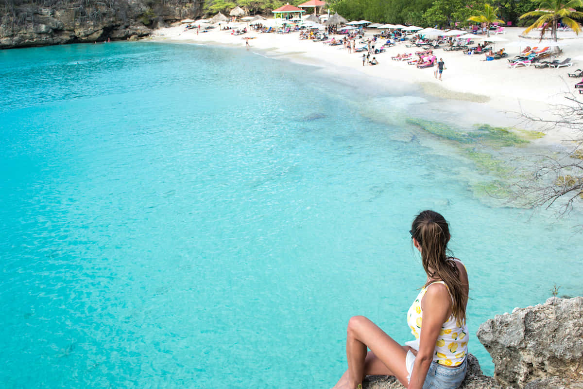 Caribbean Island 桌布 壁紙 — 一名女子坐在岩石峭壁上，俯瞰著充滿活力的碧綠海灣，沙灘上的日光浴者在白色沙灘上放鬆。