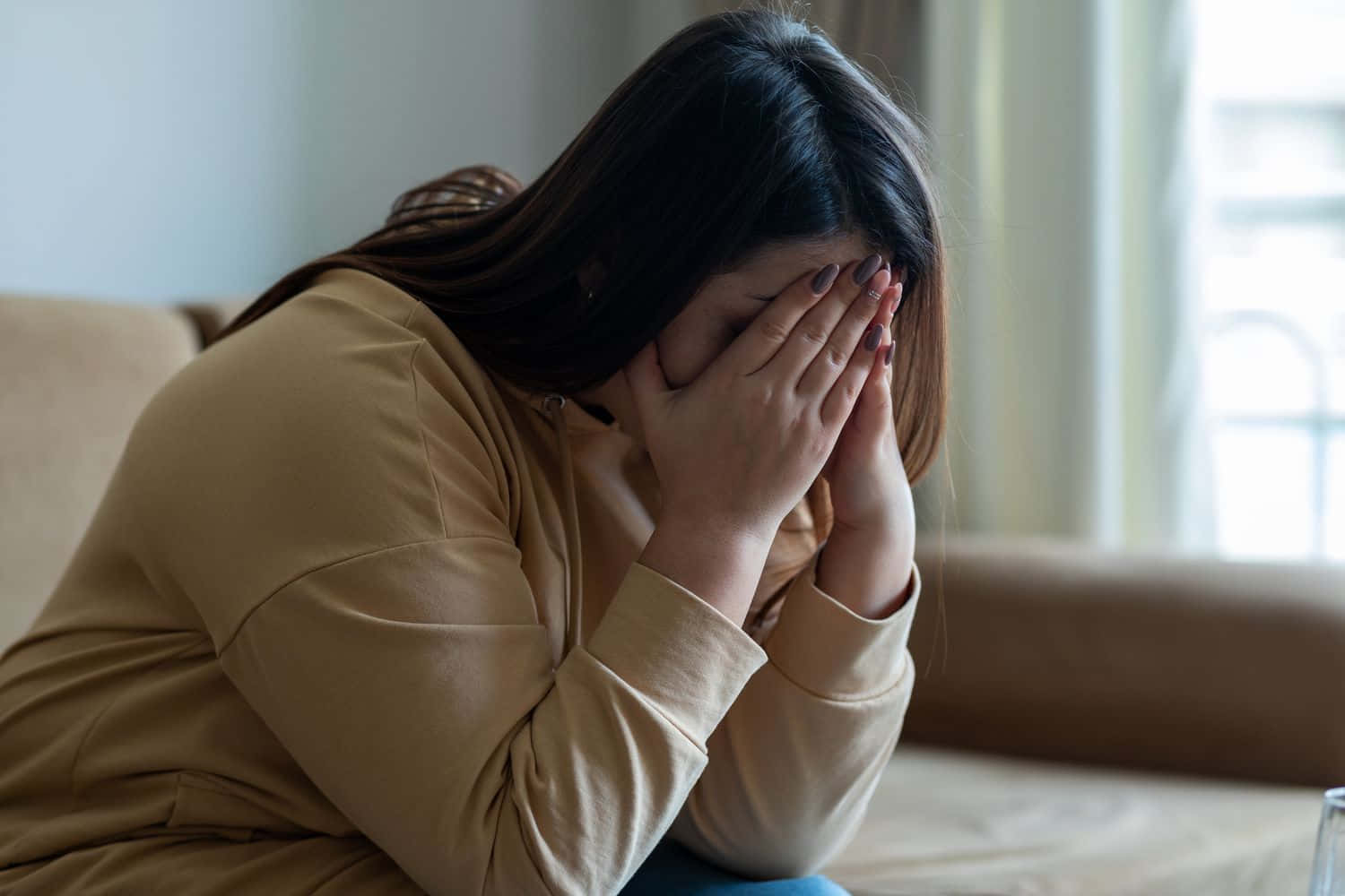 Depressed 桌布 壁紙 — 一名女子坐在沙發上，雙手抱頭，在柔和光線的房間中顯得不知所措或痛苦。