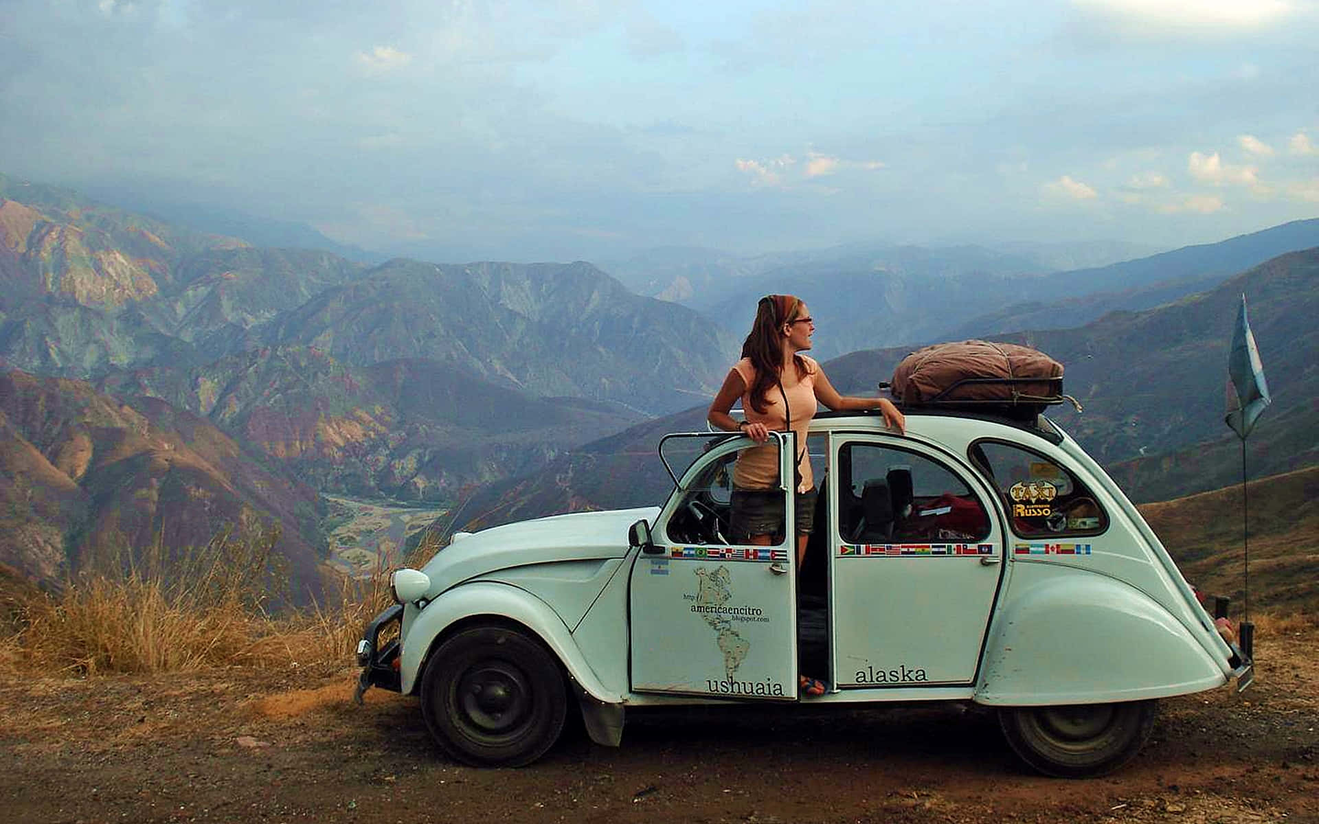 Traveler 桌布 壁紙 — 一名女子從一輛停在山上觀景台的淺藍色復古福斯金龜車中探出身子，層層疊疊、色彩斑斕的山峰延伸至朦朧的遠方。