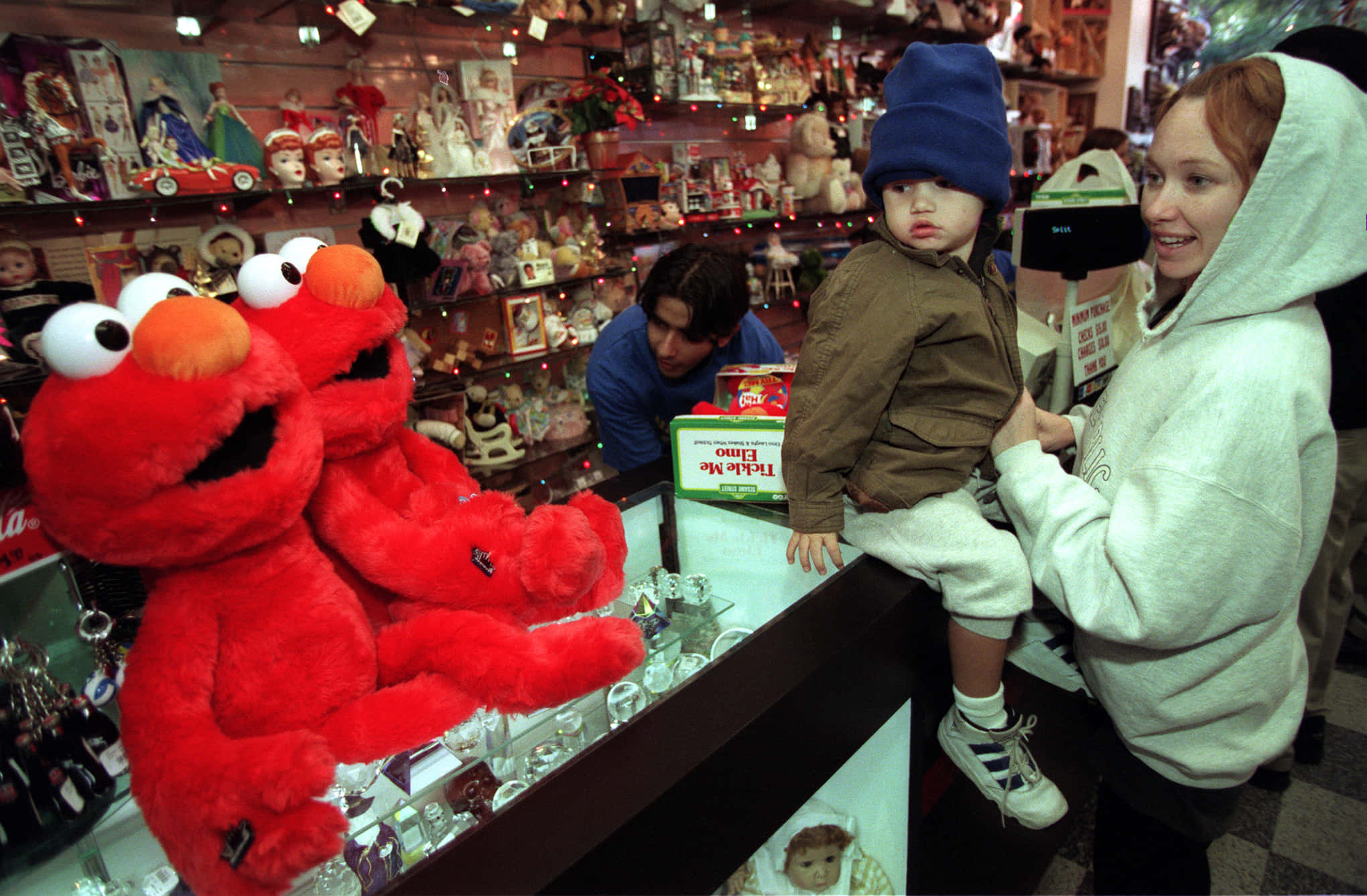 Elmo 桌布 壁紙 — 一名女子抱著一個孩子，兩人凝視著一間雜亂無章的玩具店裡展示的巨大紅色 Elmo 絨毛玩具，店內擺滿了洋娃娃和收藏品。