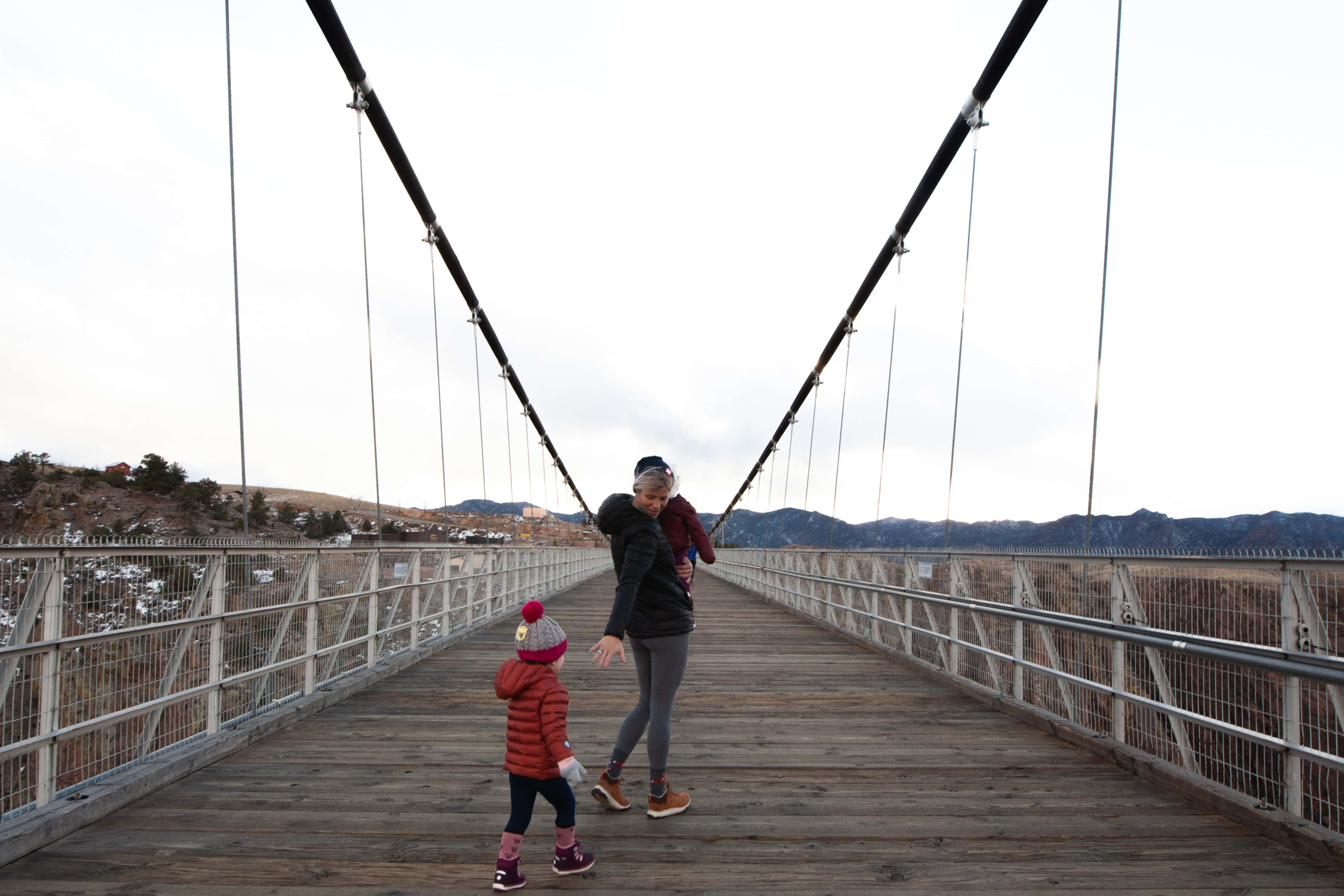 Mom 桌布 壁紙 — 一名女子抱著一個孩子，另一個孩子走在前面，在一個陰天下的山谷上方的木製吊橋上，遠處有山脈。