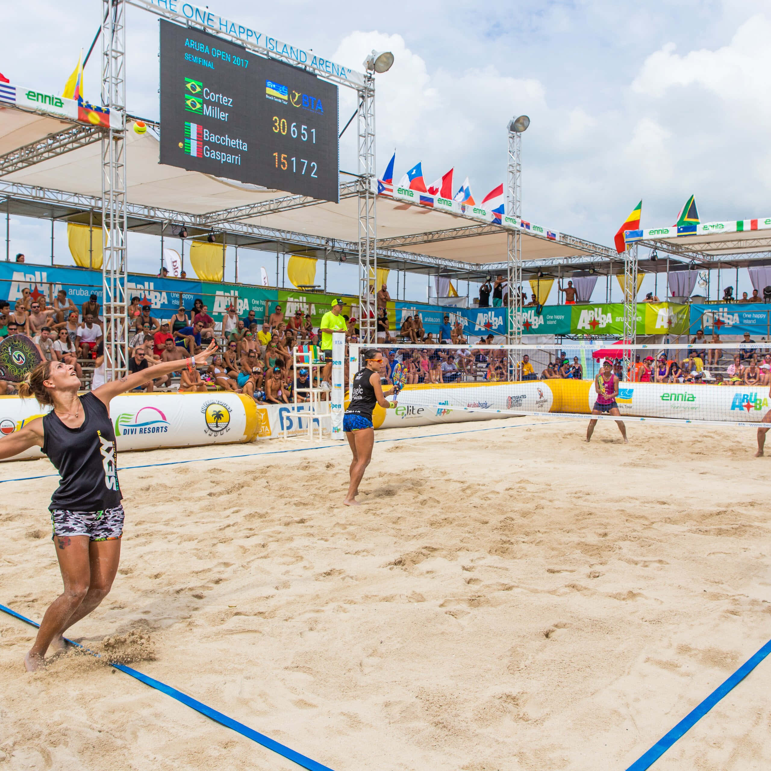 Beach Tennis 桌布 壁紙 — 一名女子沙灘排球運動員在陽光明媚的日間比賽中跳起扣球，背景有觀眾和記分板。