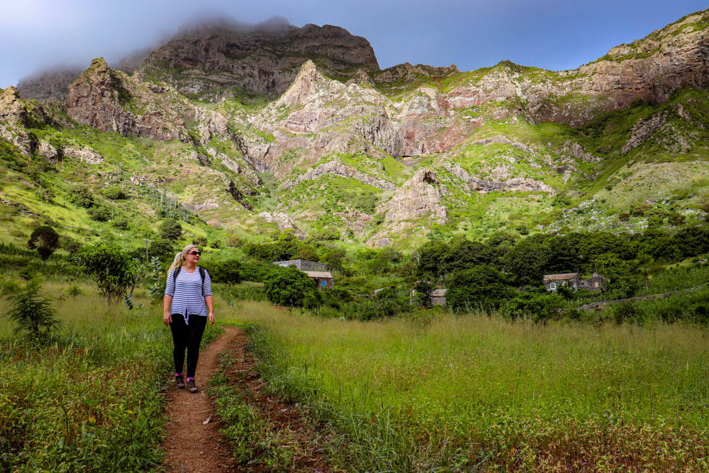 山景 桌布 壁紙 — 一名女子沿著一條泥路穿過茂密的綠色田野，背後是崎嶇的山脈，在部分多雲的天空下升起。